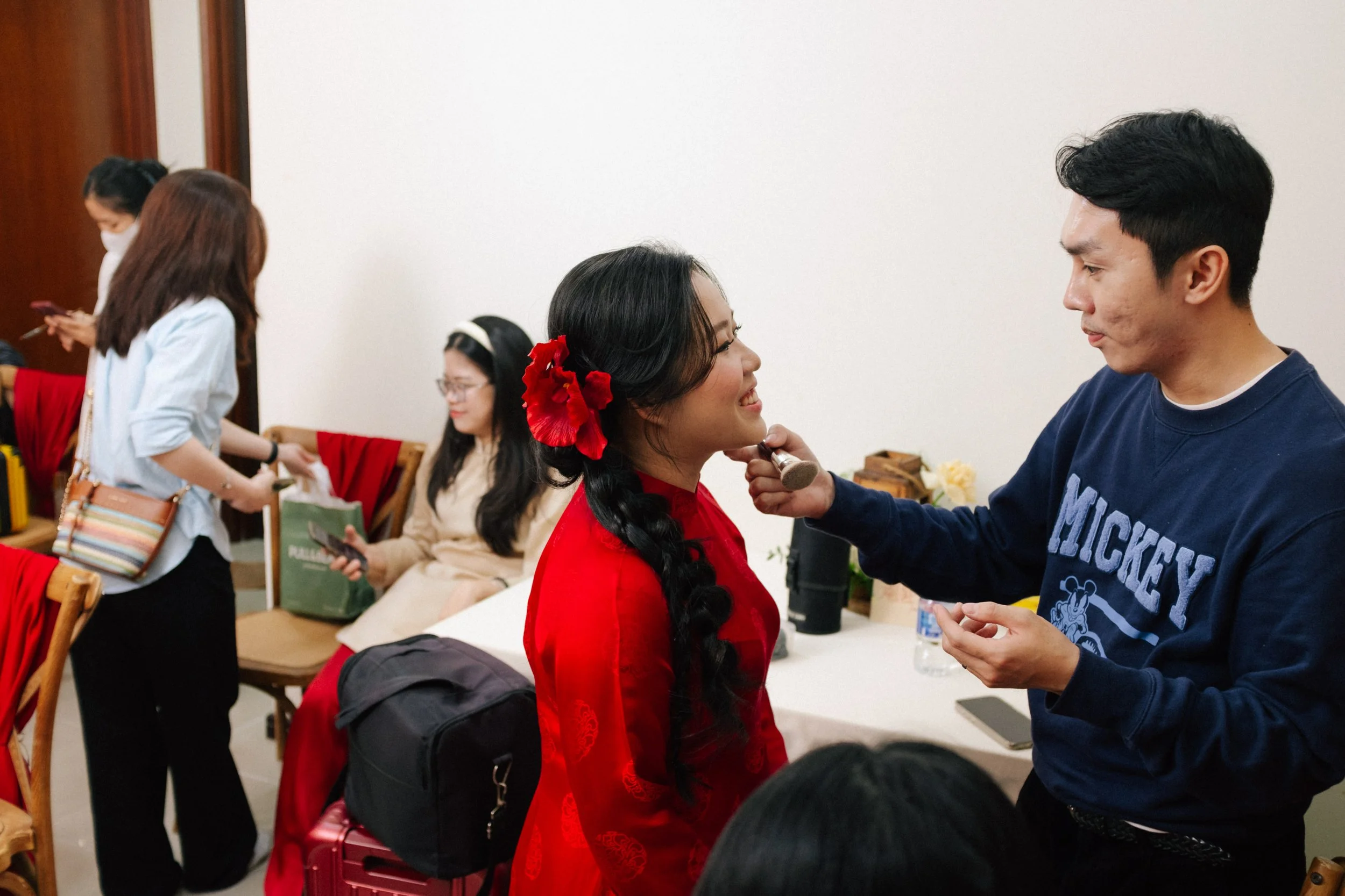 A woman in a red traditional dress getting her makeup done by a man in a blue sweatshirt. Other women are in the background, one sitting and two standing, with some using their phones.