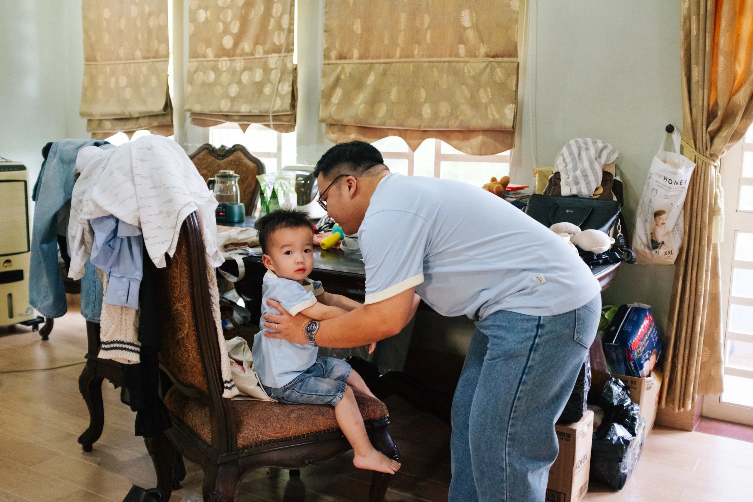 A man is holding a young boy on a chair in a cluttered room with a table, covered in clothes, bags, and various items. The boy looks at the camera, and the man is leaning in close to him.