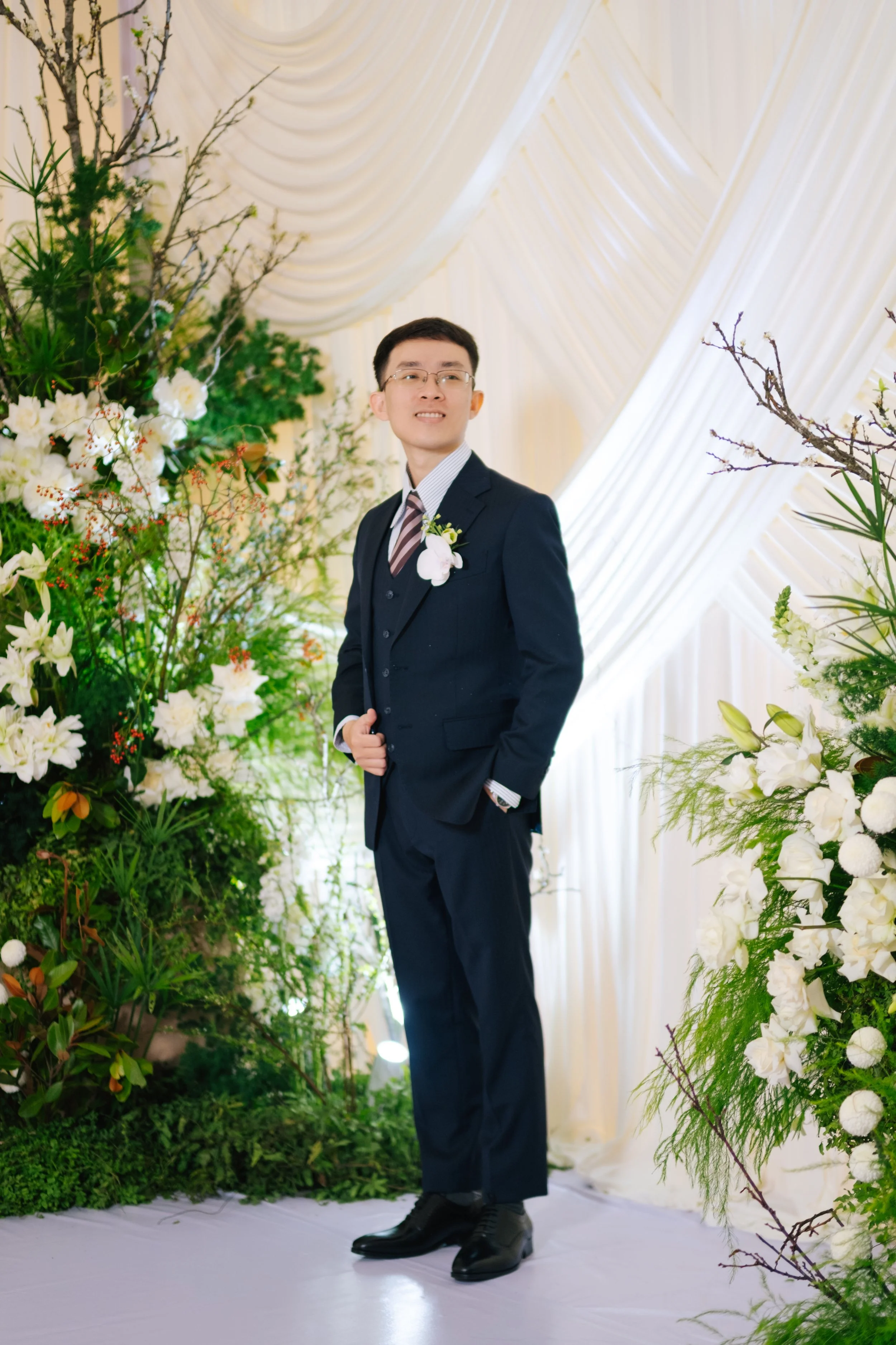 A young man in a dark suit with a striped tie and glasses, standing in a floral decorated space with white drapes, smiling and looking slightly to the side.