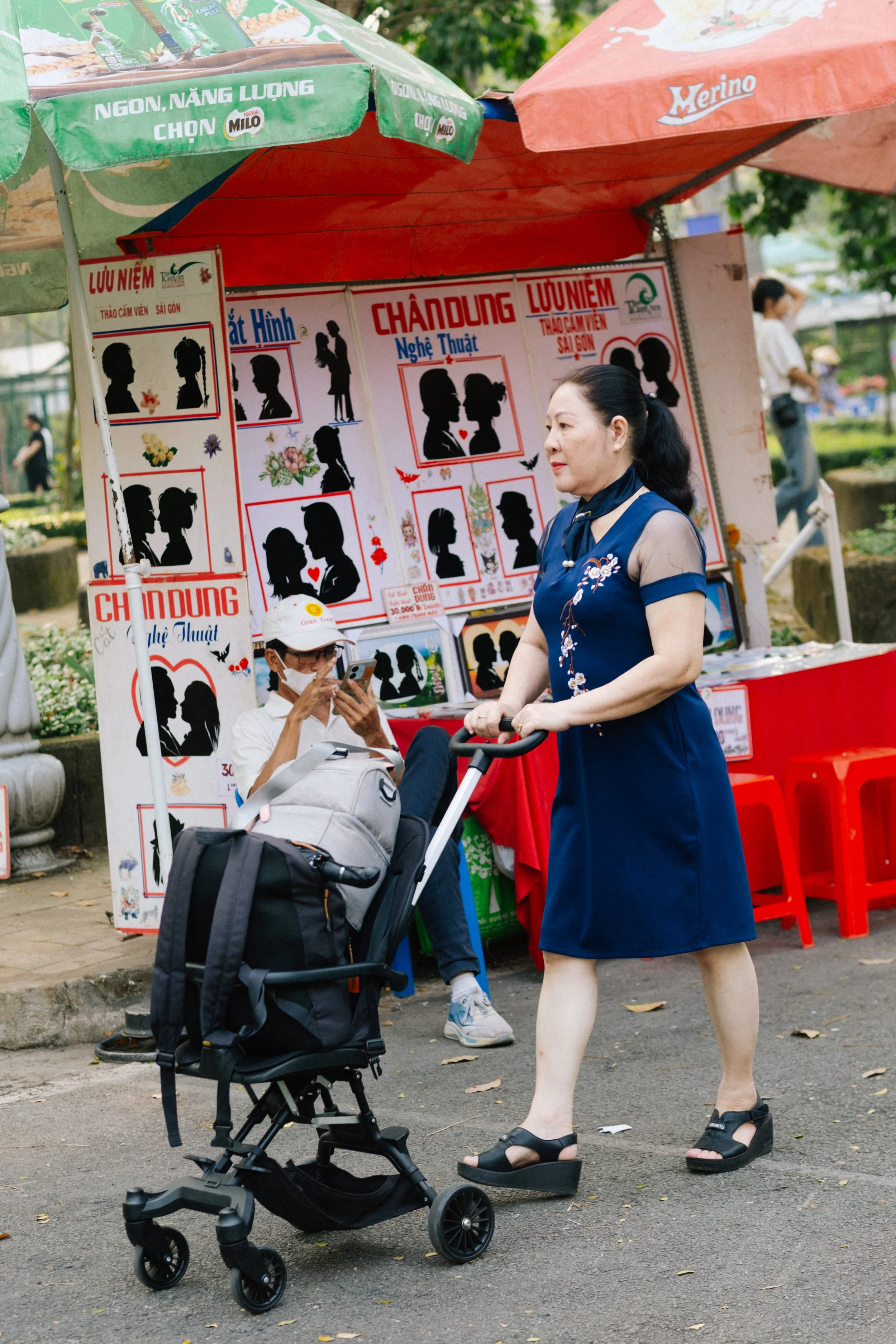 A woman with dark hair in a ponytail wearing a blue dress and black sandals, pushing a stroller on a sidewalk in a park. Behind her, there is a woman sitting and looking at her phone near a booth with posters and silhouettes of couples, indicating it