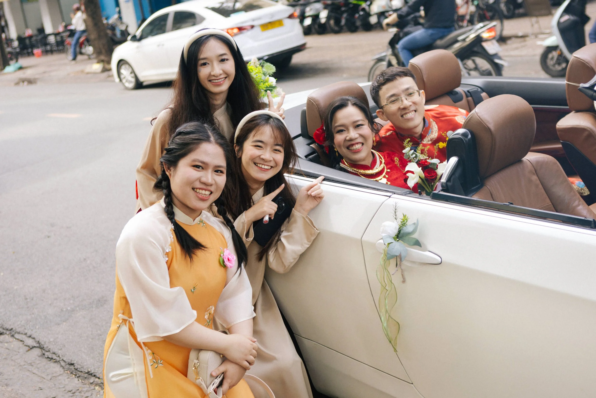 A group of five young people celebrating a wedding, with some in traditional attire and one couple in a festive car with floral decorations, smiling and posing together on a city street.