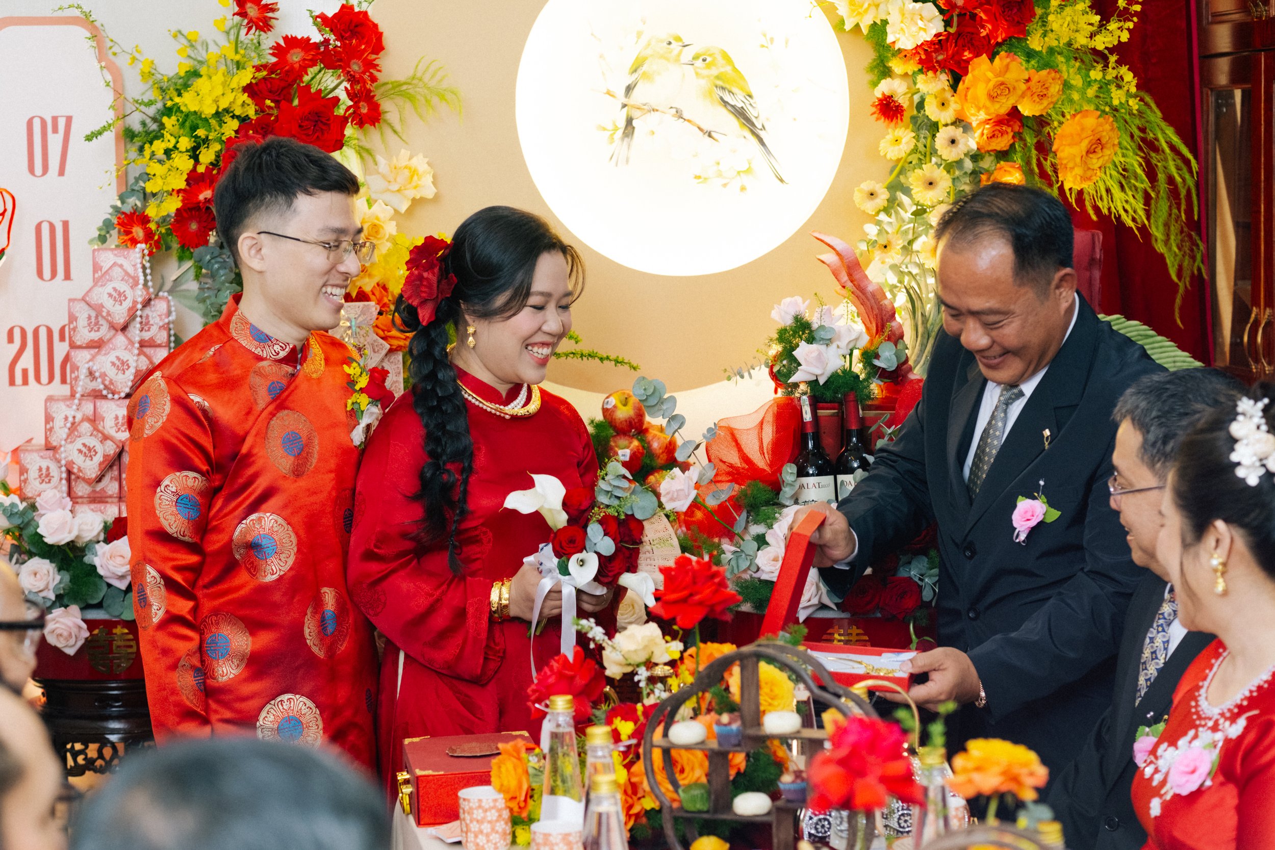 A Vietnamese wedding ceremony with the bride and groom dressed in traditional red attire, exchanging gifts, surrounded by floral decorations and guests.