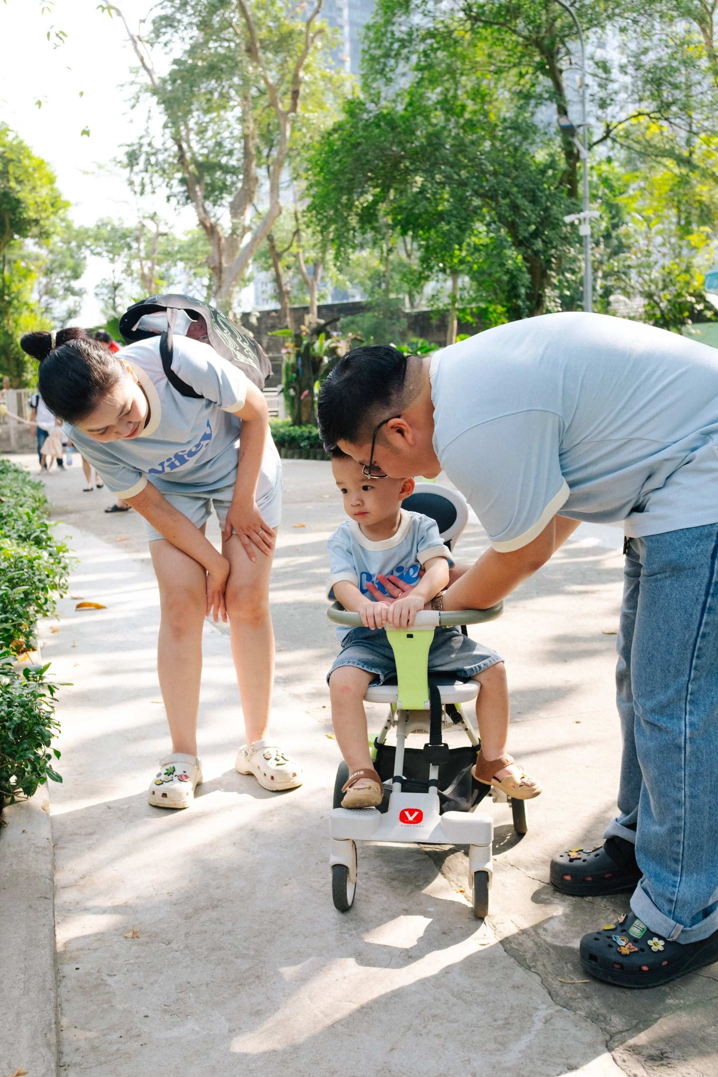A young boy sitting in a stroller outdoors, with two adults leaning over him and engaging with him on a sunny day in a park.
