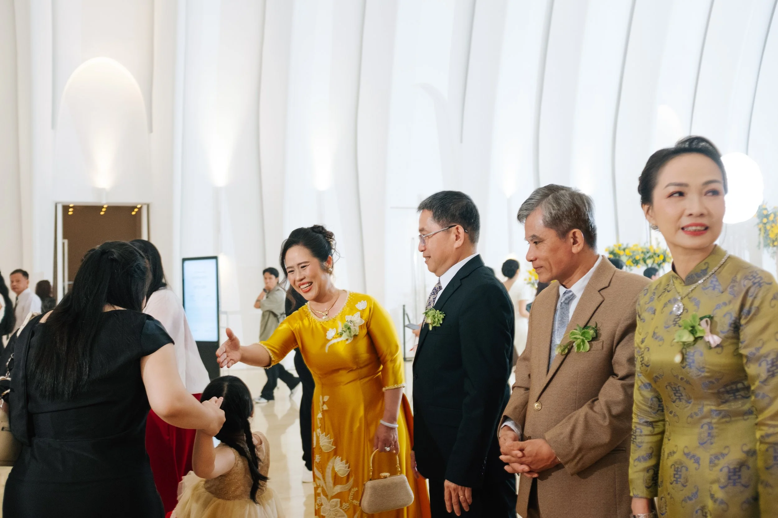Group of people at a formal event greeting each other, with women in traditional Asian attire and men in suits, inside a bright, modern venue.