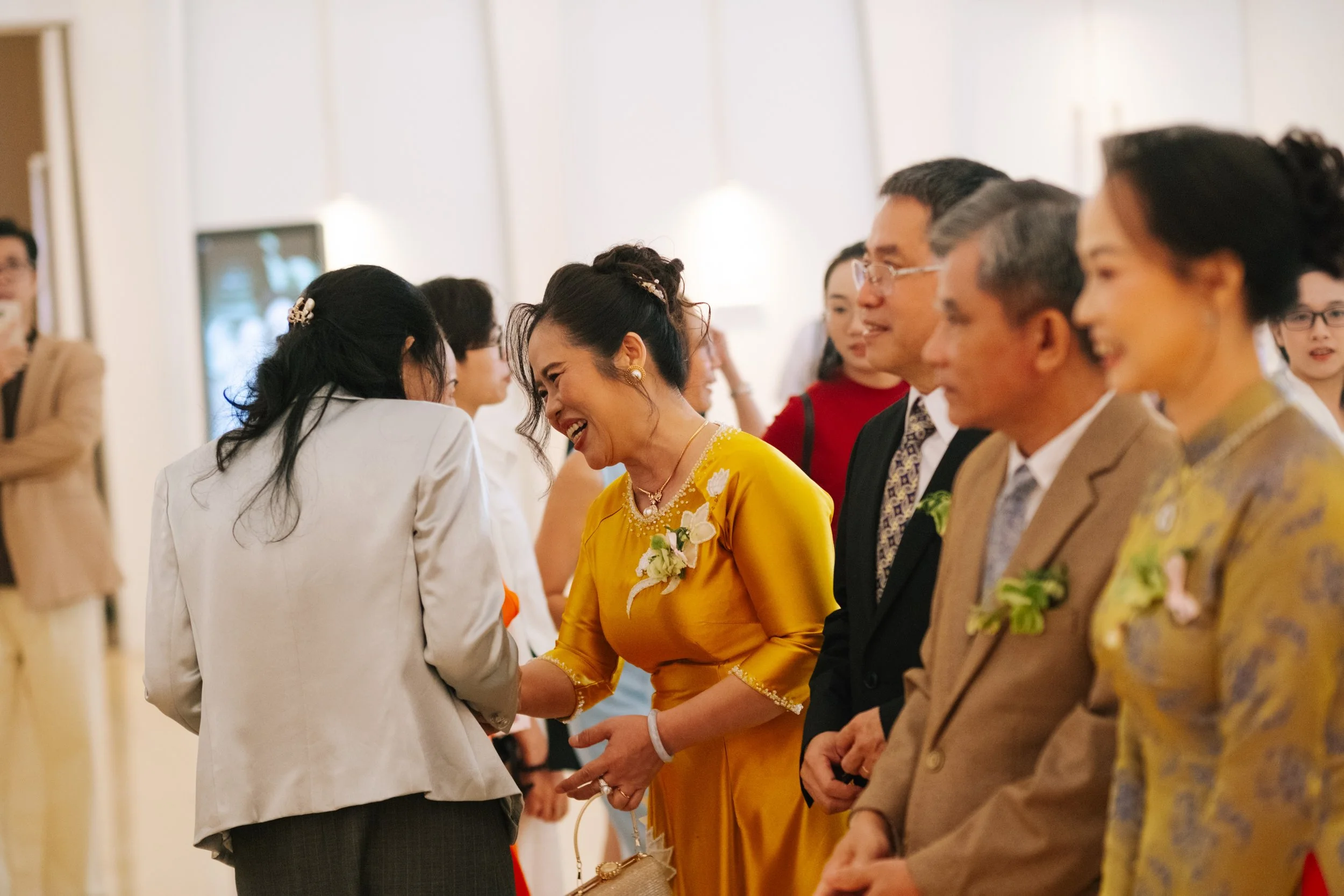 People laughing and smiling at a formal event, with a woman in a yellow dress and a woman in a white jacket engaging in conversation.