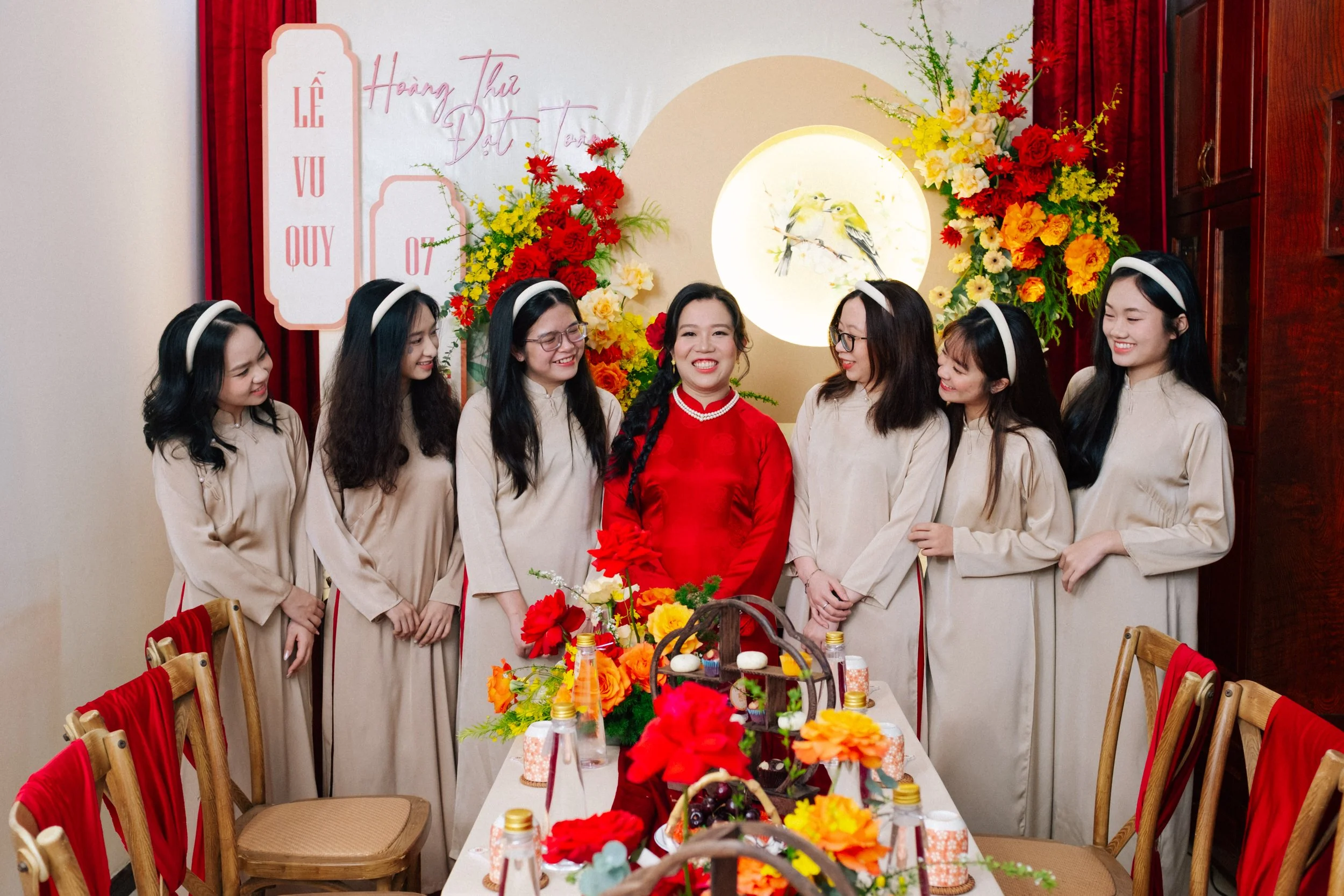 A woman in a red traditional dress standing in the middle of six women in beige áo dài, all wearing white headbands, in a decorated room with floral arrangements and a table set with food and drinks.