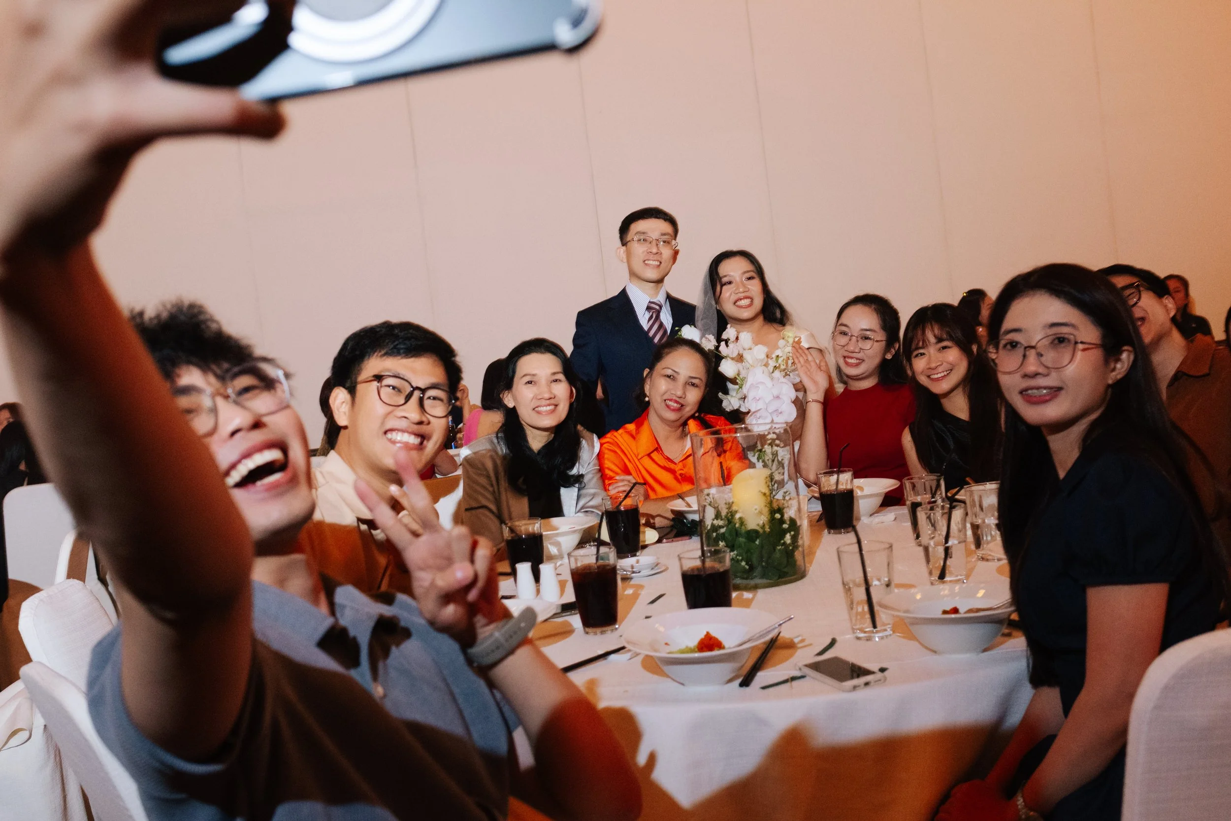 Group of people at a celebration or wedding, smiling, taking a selfie, seated around a table with food and drinks