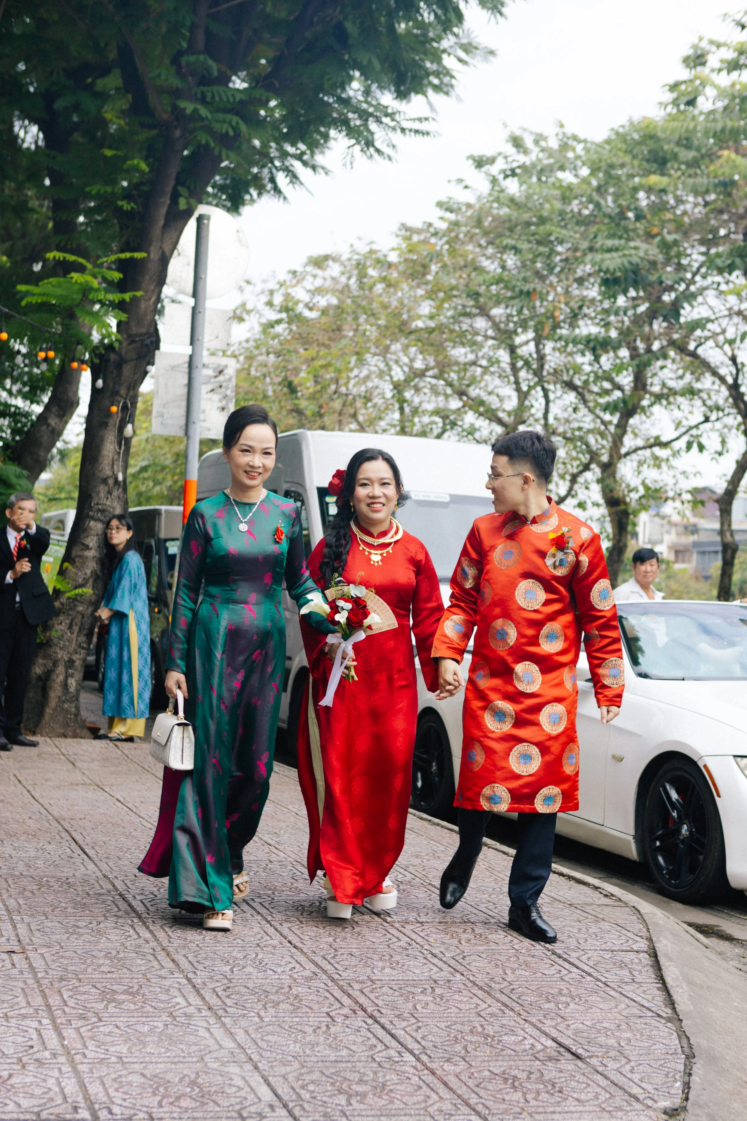A group of people dressed in traditional Vietnamese áo dài walking outdoors during a celebration, with a white car, trees, and other guests in the background.