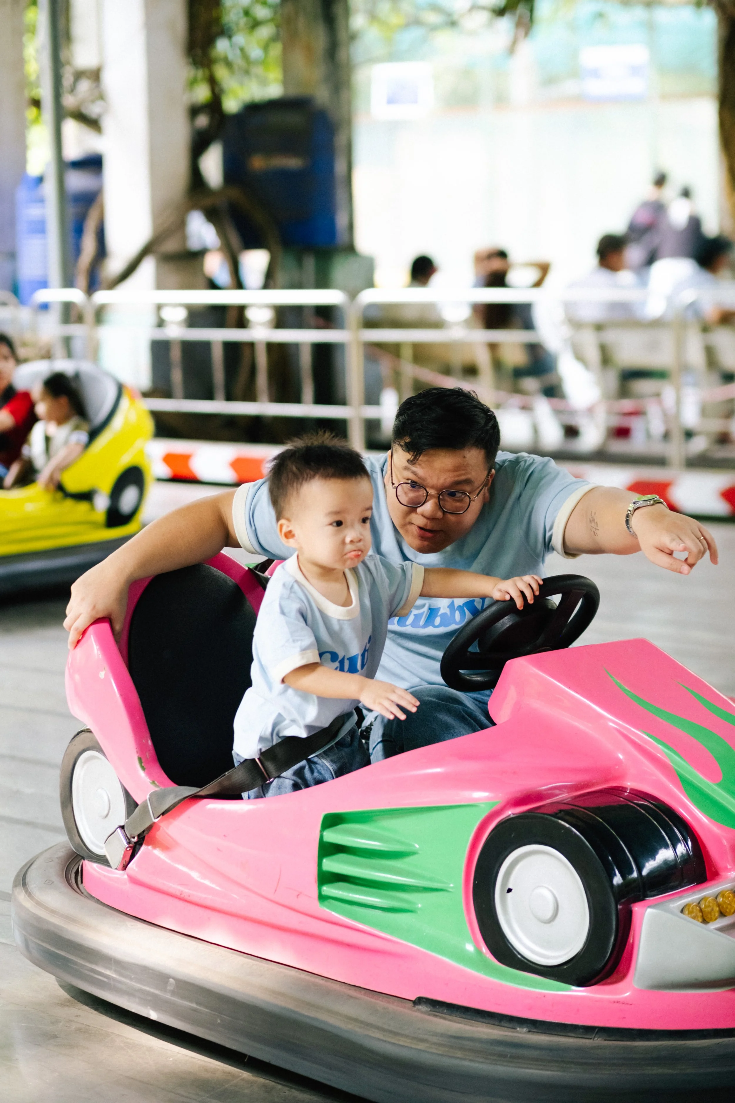 A man and a young boy riding in a pink bumper car at an amusement park or fairground.
