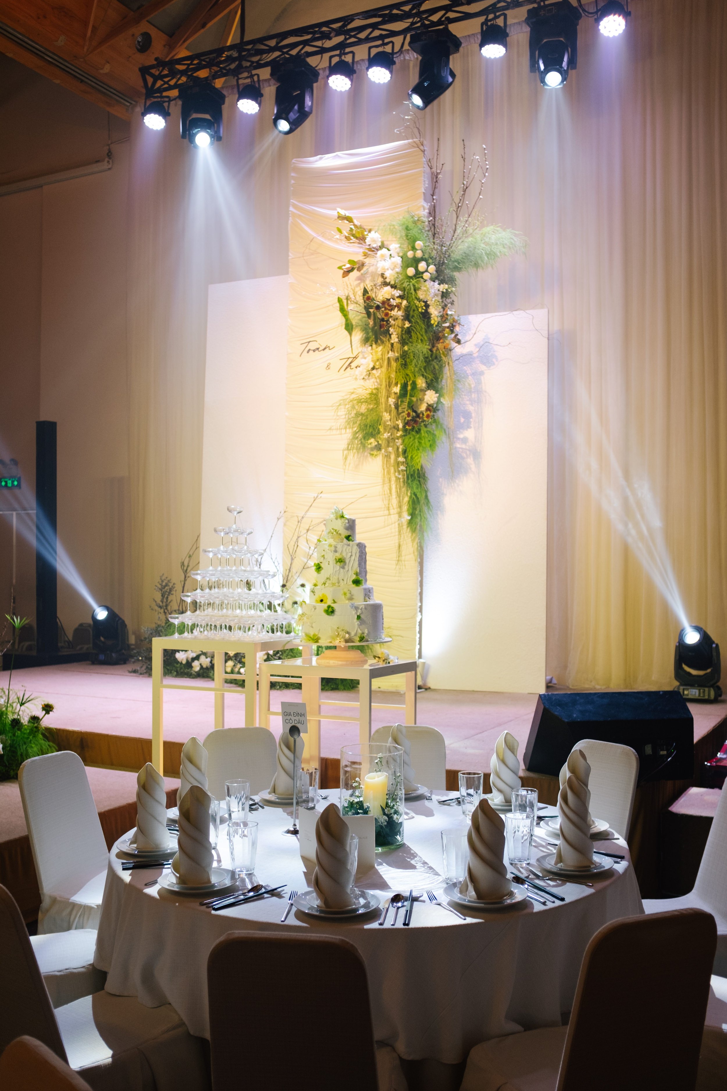 Wedding banquet table in front of a decorated stage with a tiered cake and champagne glasses arrangement, under stage lighting.