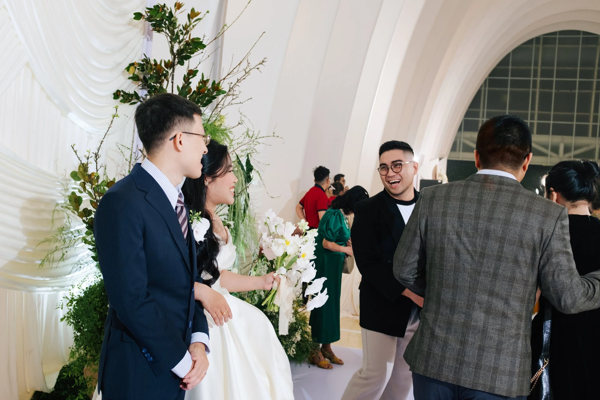People dressed in formal attire socializing at a wedding reception, with a bride holding a bouquet of flowers and standing next to a groom, surrounded by guests in an elegant venue with floral decorations.