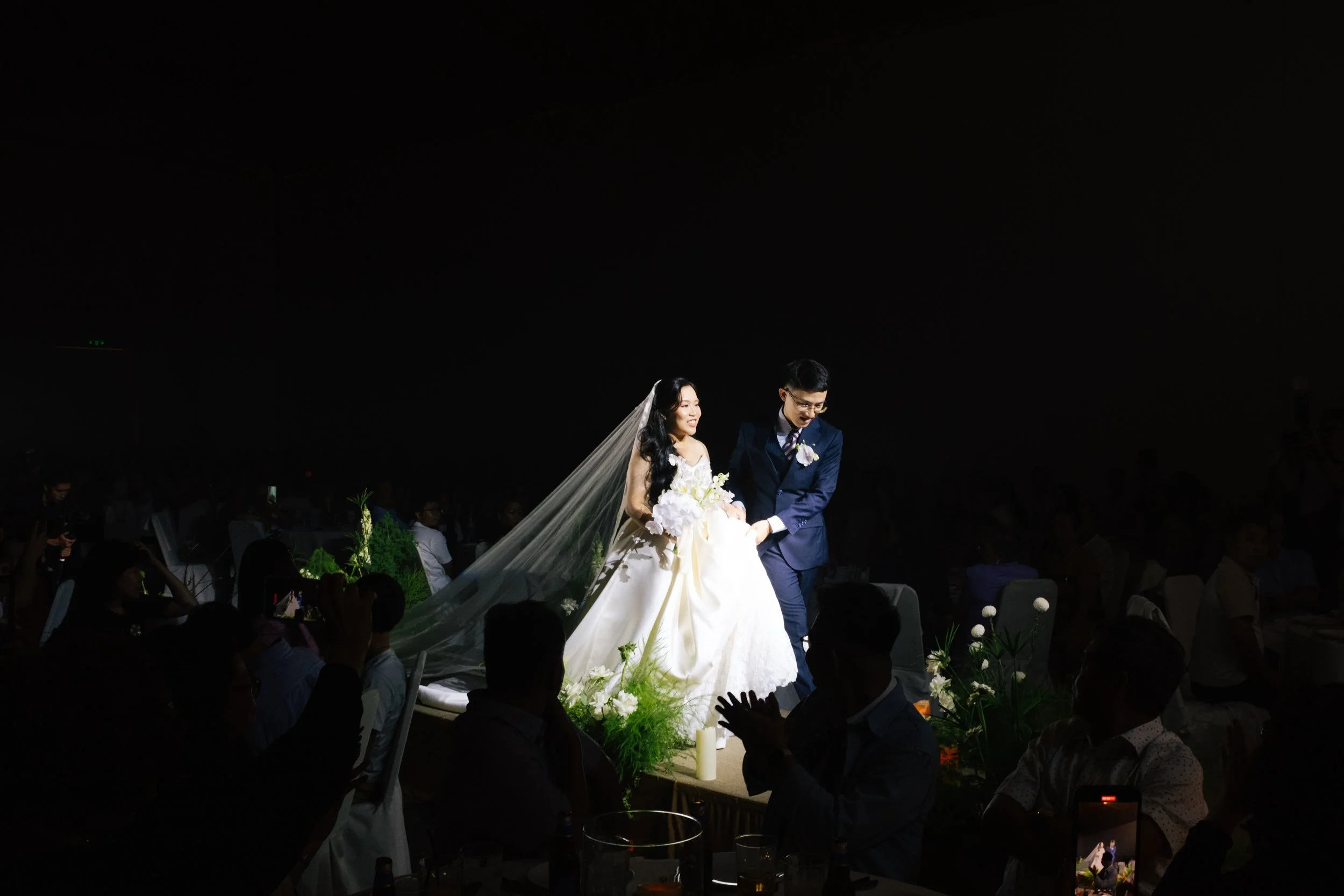 A bride and groom walking down the aisle during their wedding ceremony, surrounded by guests in a dimly lit room.