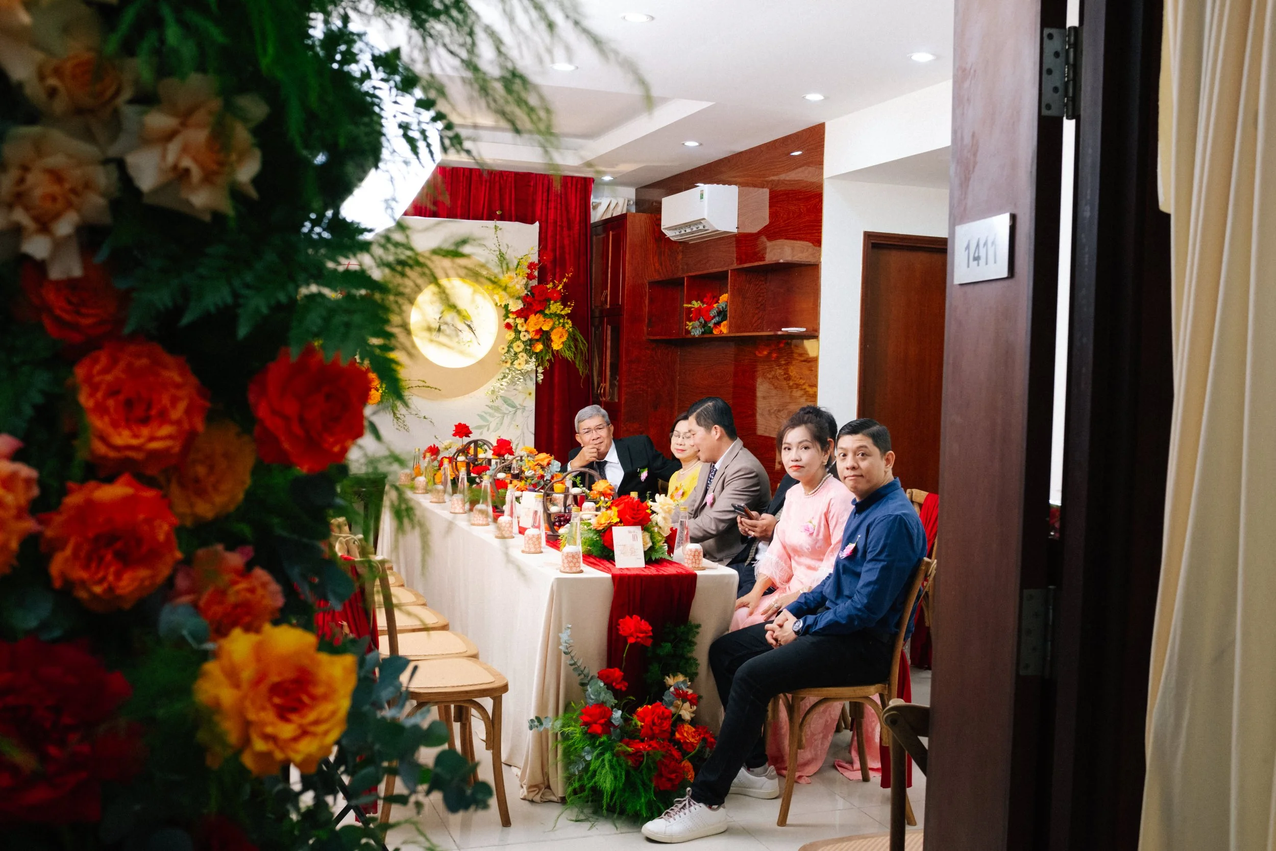People sitting at a decorated banquet table in a room with wooden paneling, red curtains, and floral arrangements.