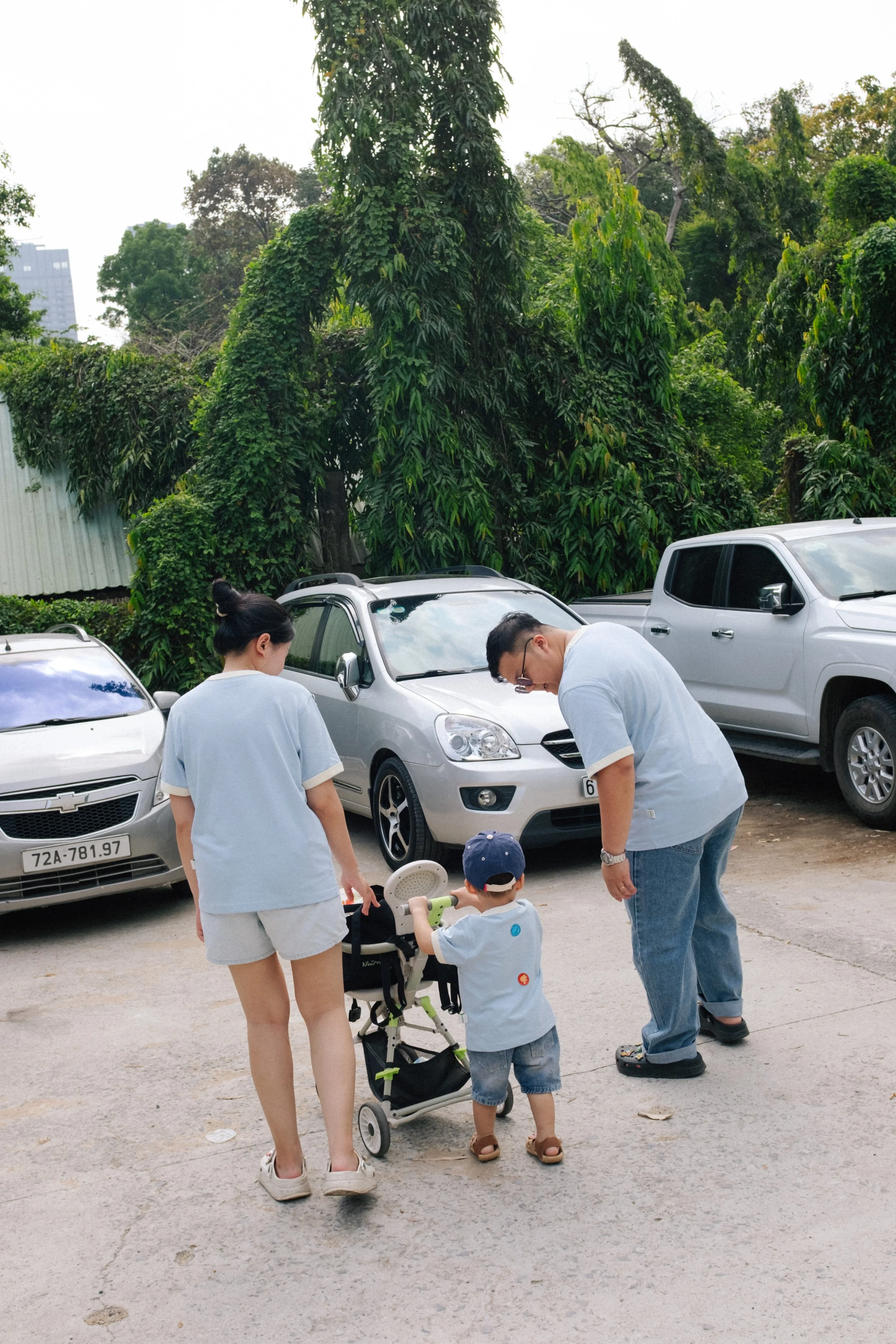 A family of three, including a woman, man, and young boy, standing near parked cars in a driveway with greenery and trees in the background.