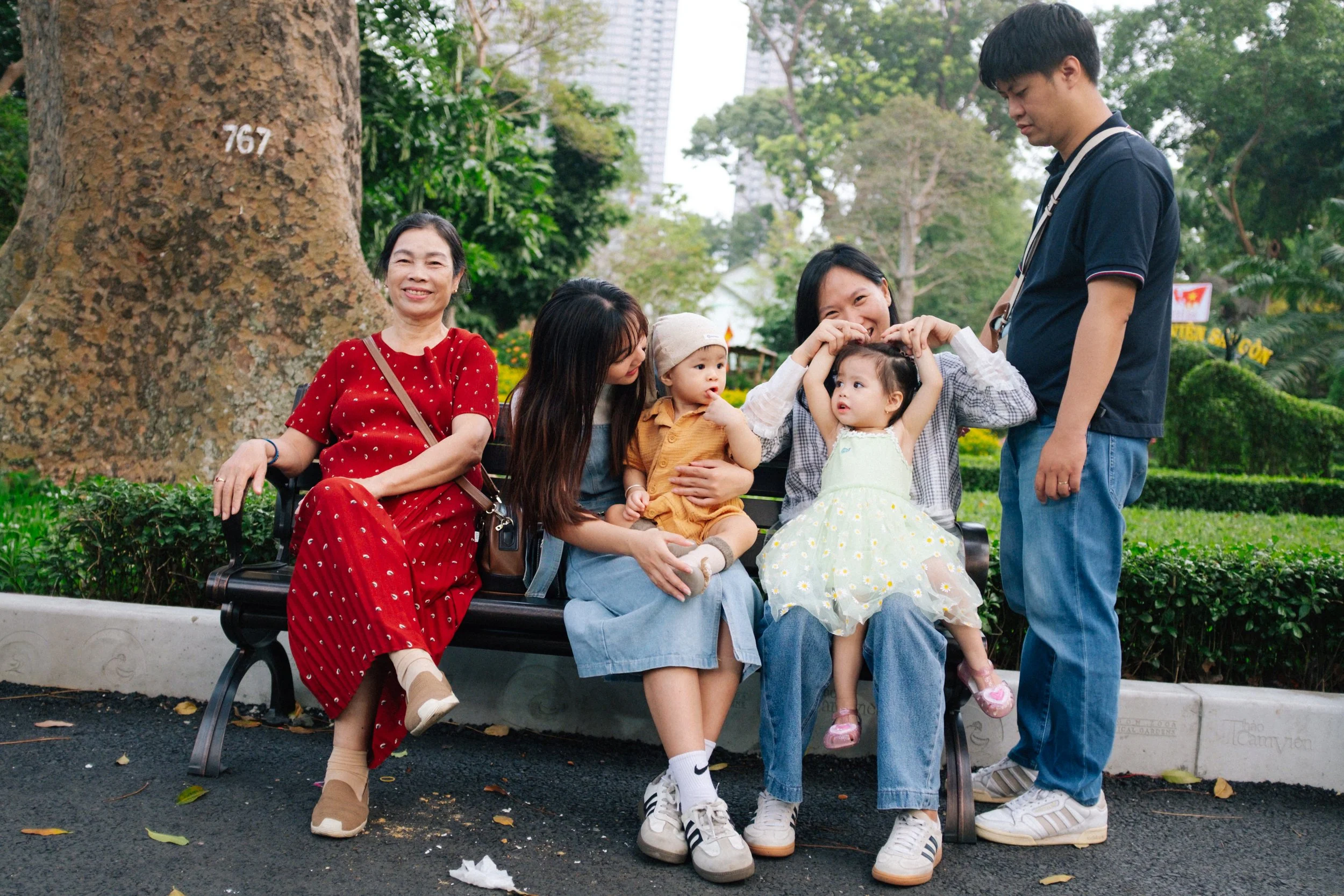 A multigenerational Asian family sitting on a park bench with lush greenery and trees in the background. The group includes an elderly woman in a red dress, a young woman with two children, and a young man. The children are making heart shapes with t