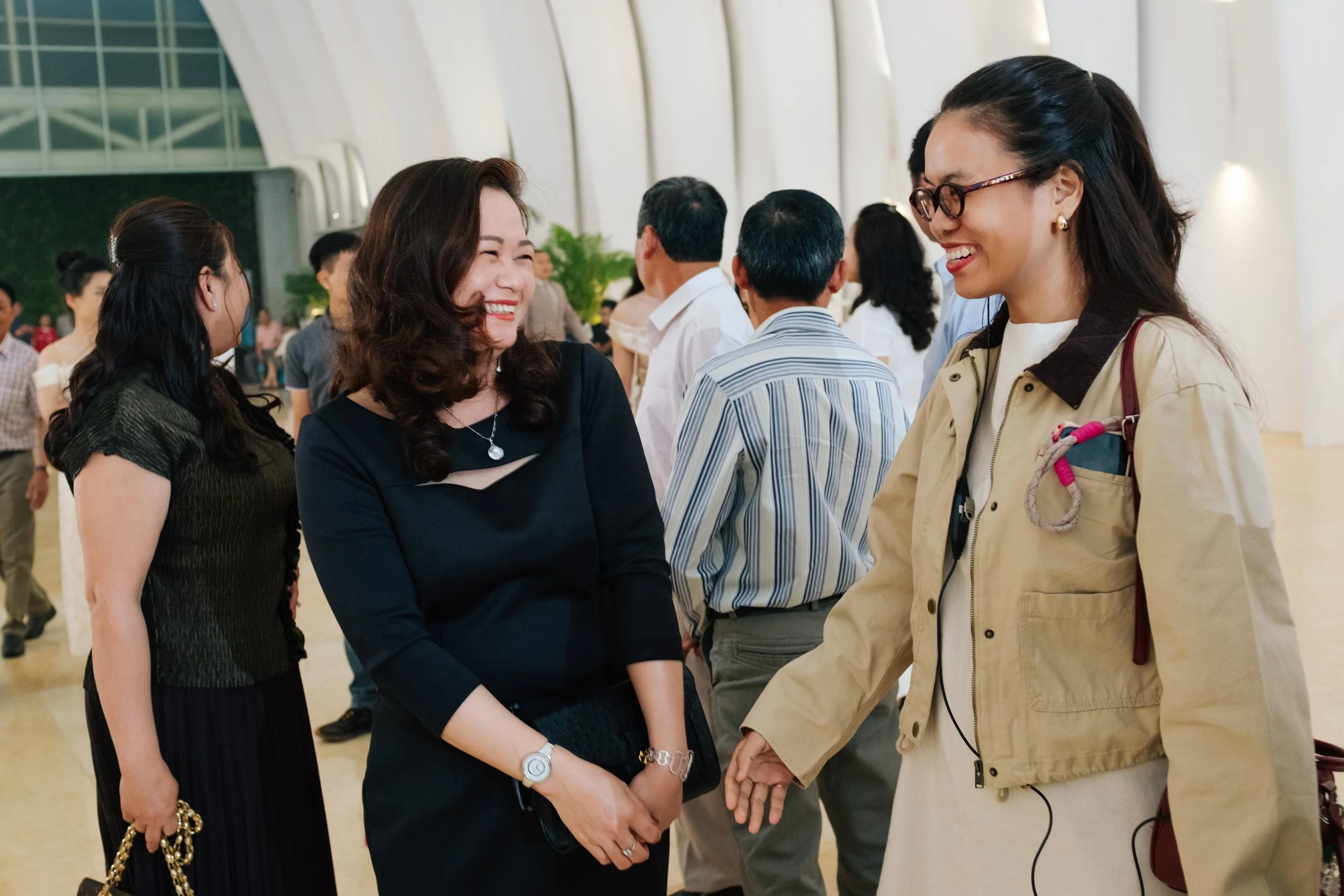 Two women smiling and talking at a social event in a large, indoor venue with many people in the background.