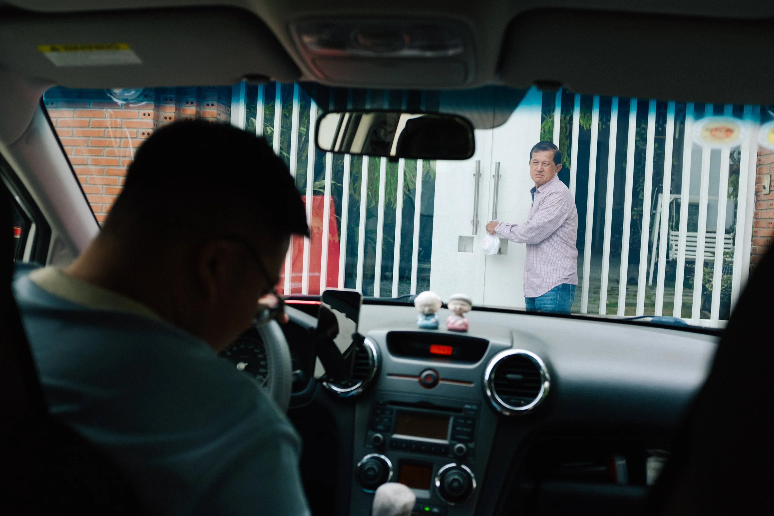 A man sitting in a vehicle is looking at another man outside near a gate, holding a plastic bag. The vehicle's dashboard has two small figurines on it, and the view is through the front windshield.