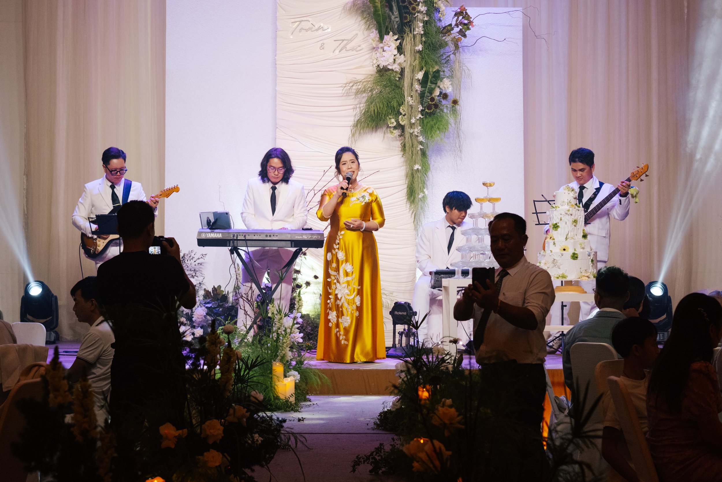 A live band performs on stage at a wedding or event, with a woman in a yellow dress singing, surrounded by musicians and decorated with flowers and candles.