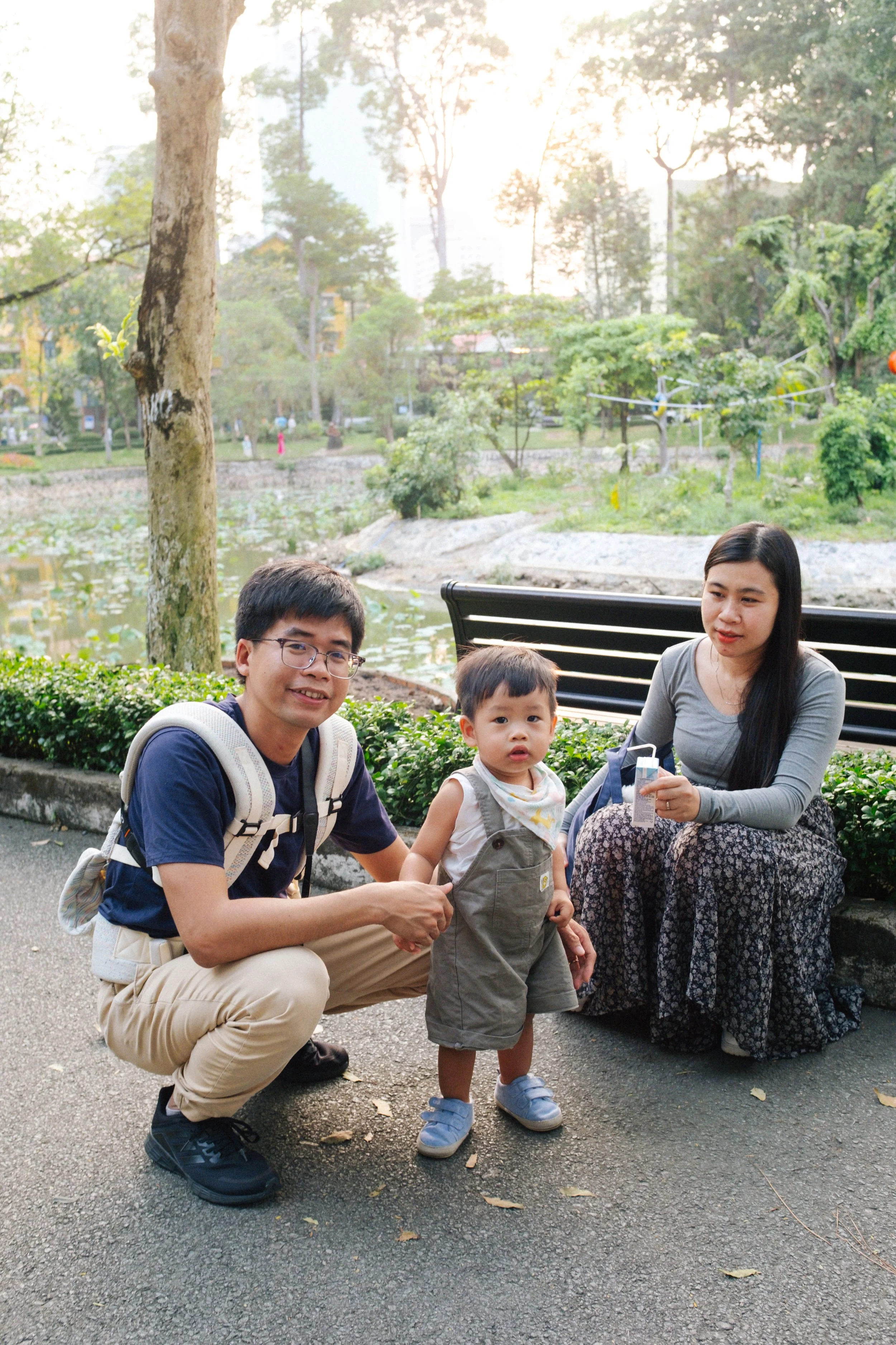 A family of three outdoors in a park with trees, a pond, and bench in the background. The man is crouching and holding the child's hand, woman is sitting on a bench, and child standing between them.