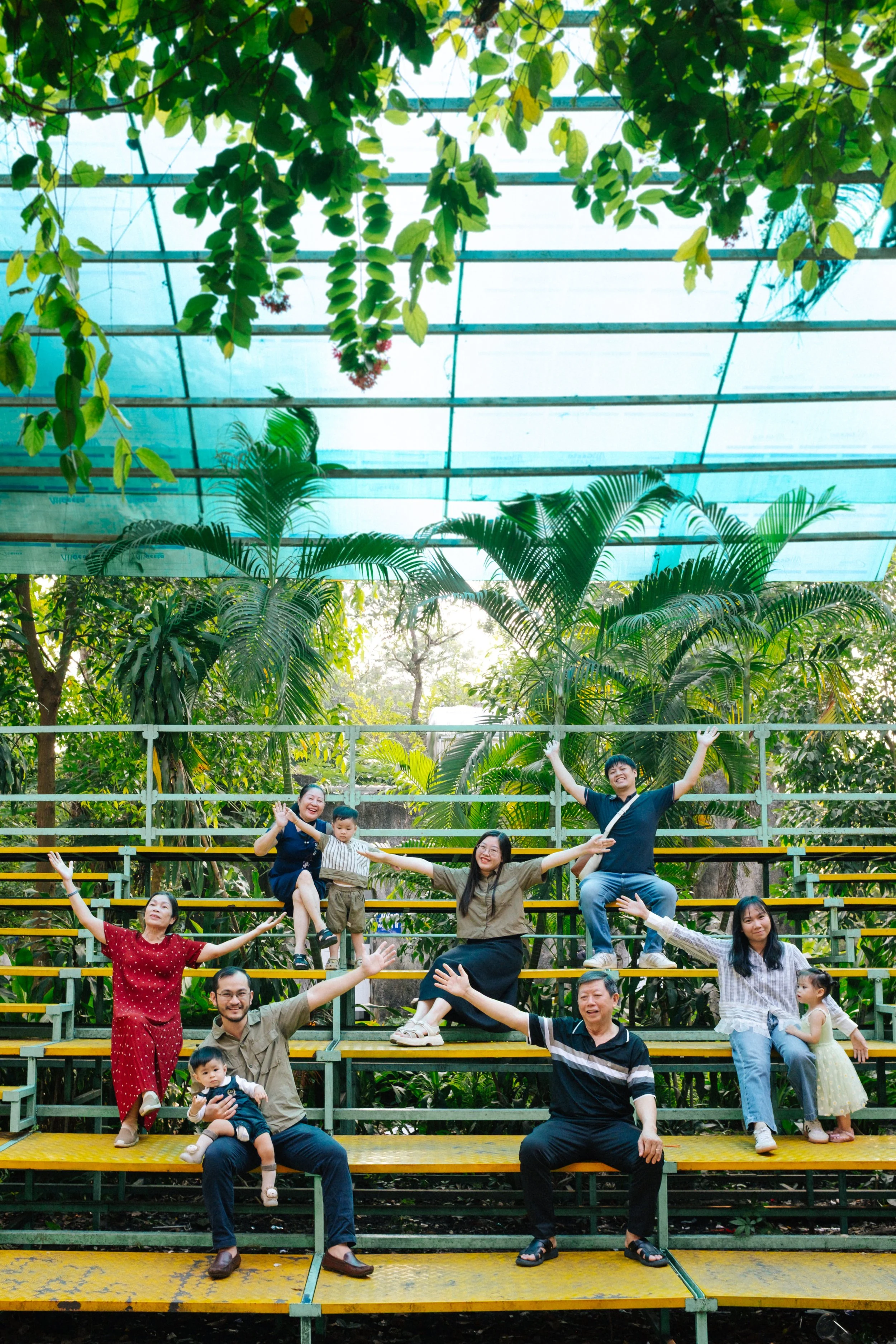 A group of ten people, including children, sitting and standing on yellow and green outdoor bleachers, with lush greenery and trees in the background, and a glass ceiling overhead.