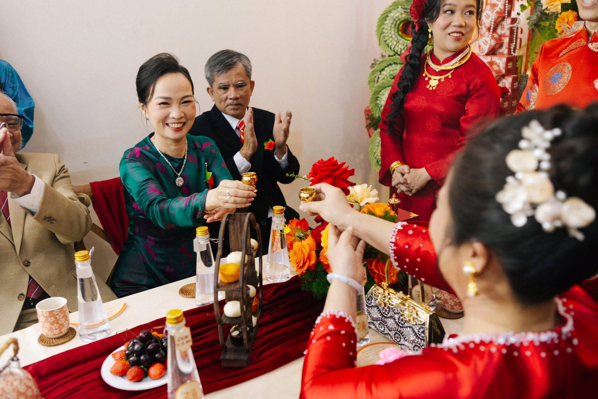 A traditional wedding tea ceremony with women in red and women in green dresses exchanging teacups, surrounded by flowers and refreshments.