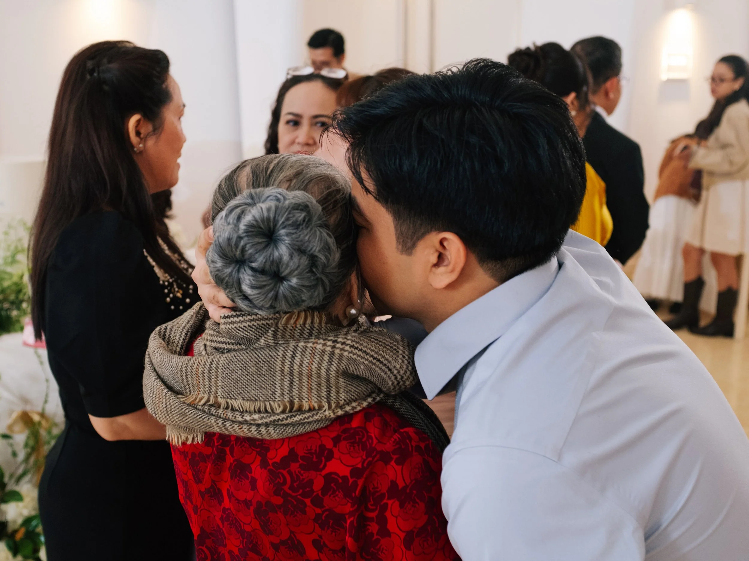 A young man kissing an elderly woman on the cheek at a gathering, with other people talking in the background.