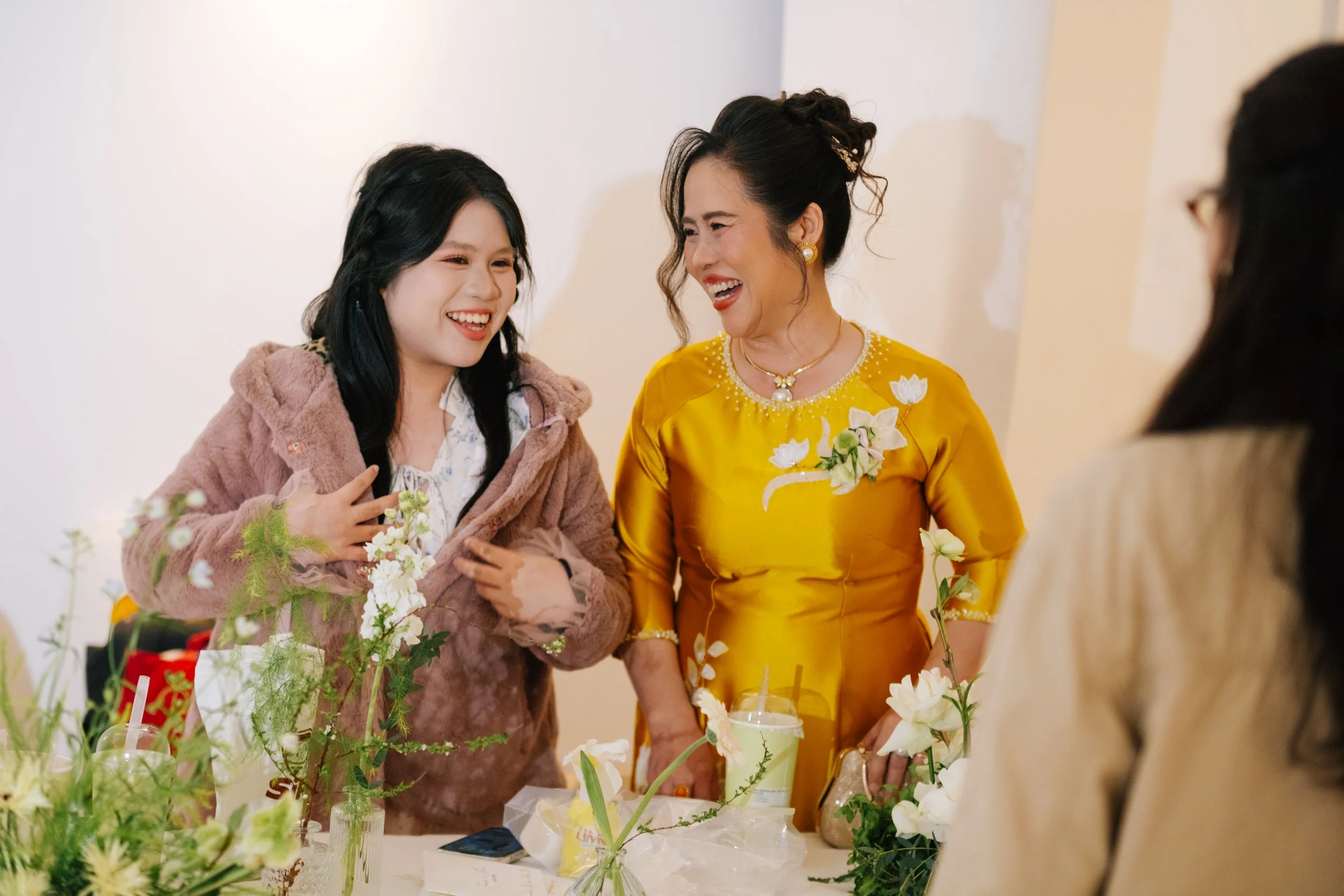 Four women smiling and laughing together at a celebration, with flowers and drinks on the table.