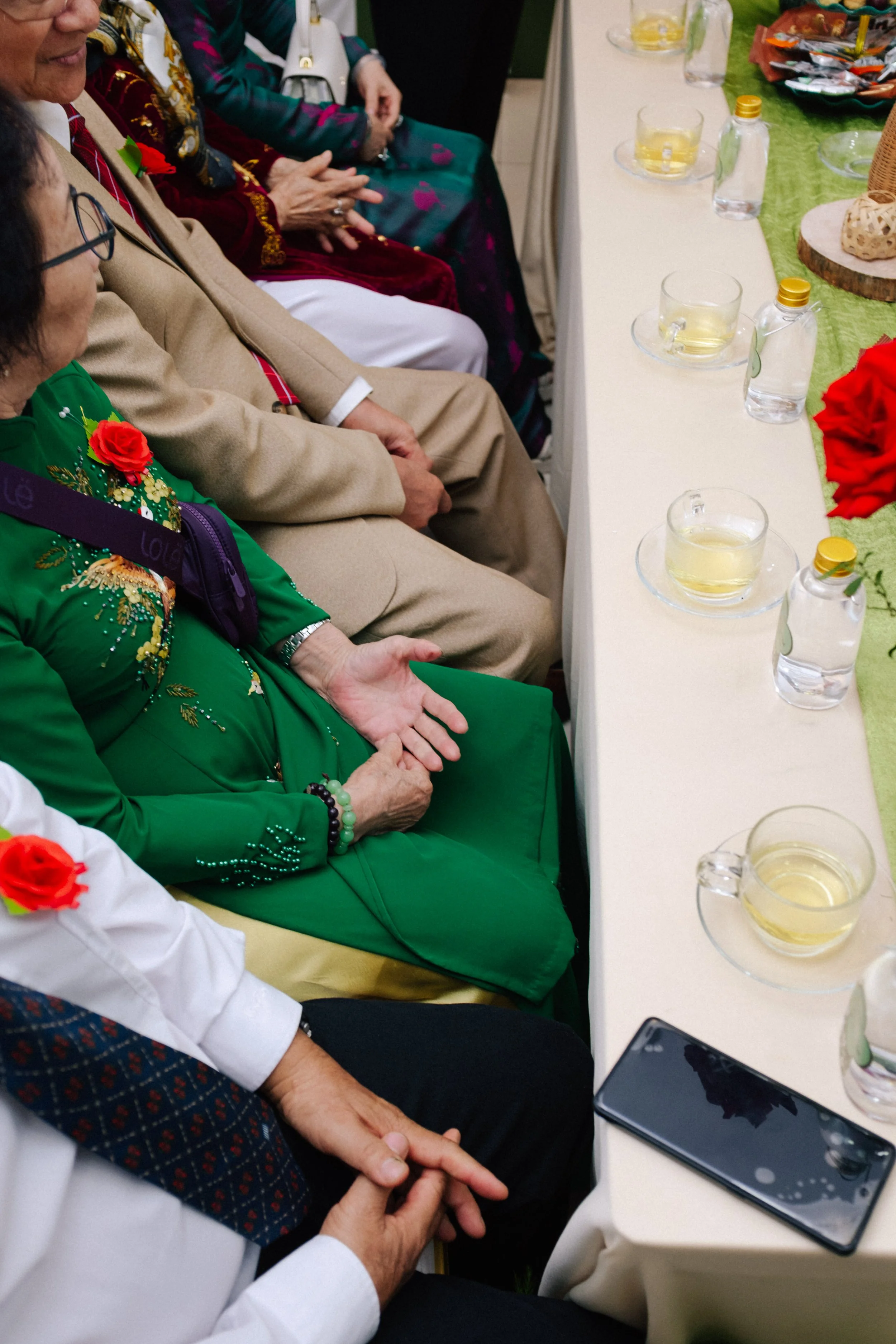 People dressed in formal attire sitting at a banquet table, with glasses and bottles of water and tea, and a smartphone on the table.