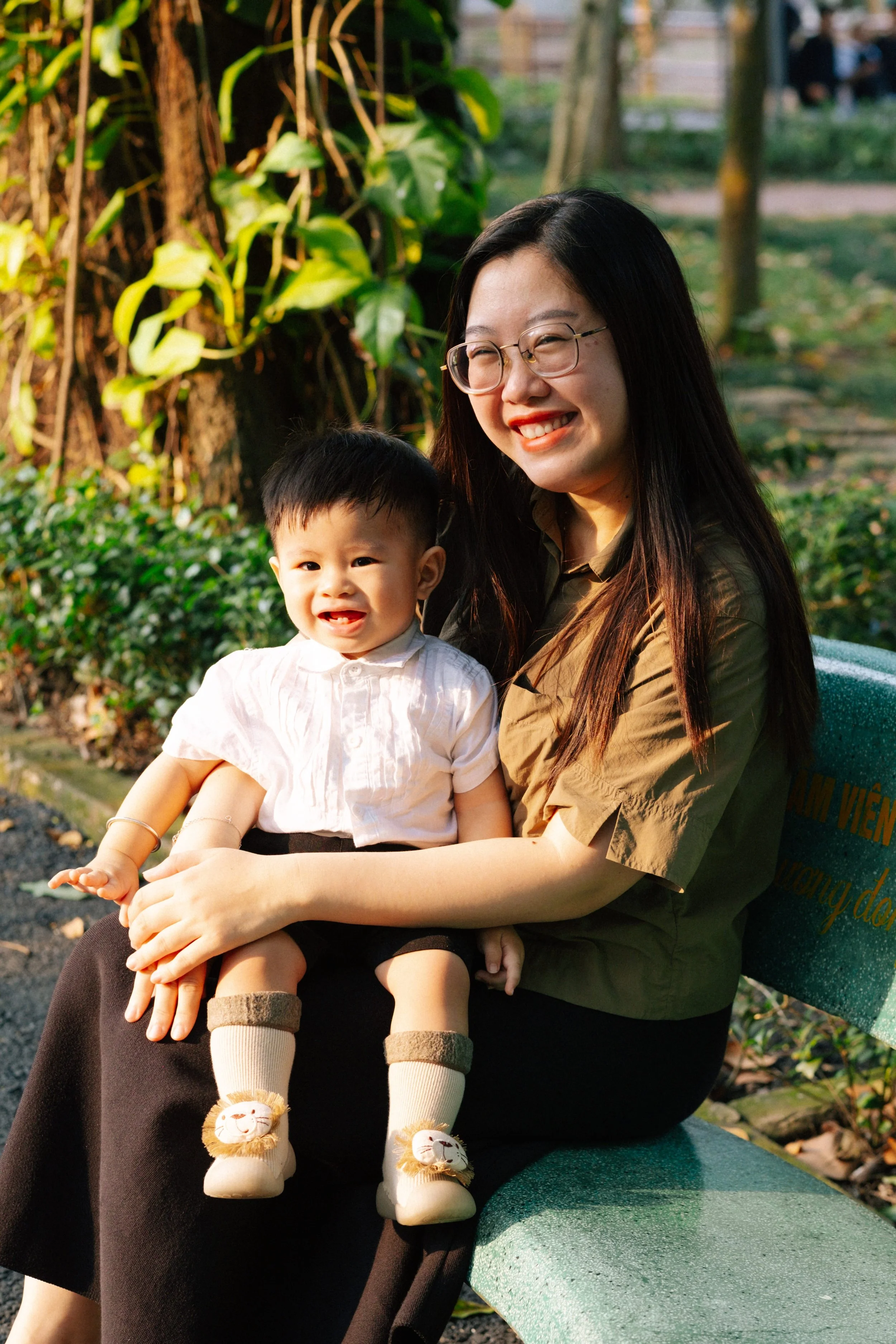 A woman and a young boy sitting on a park bench, smiling, with greenery in the background.
