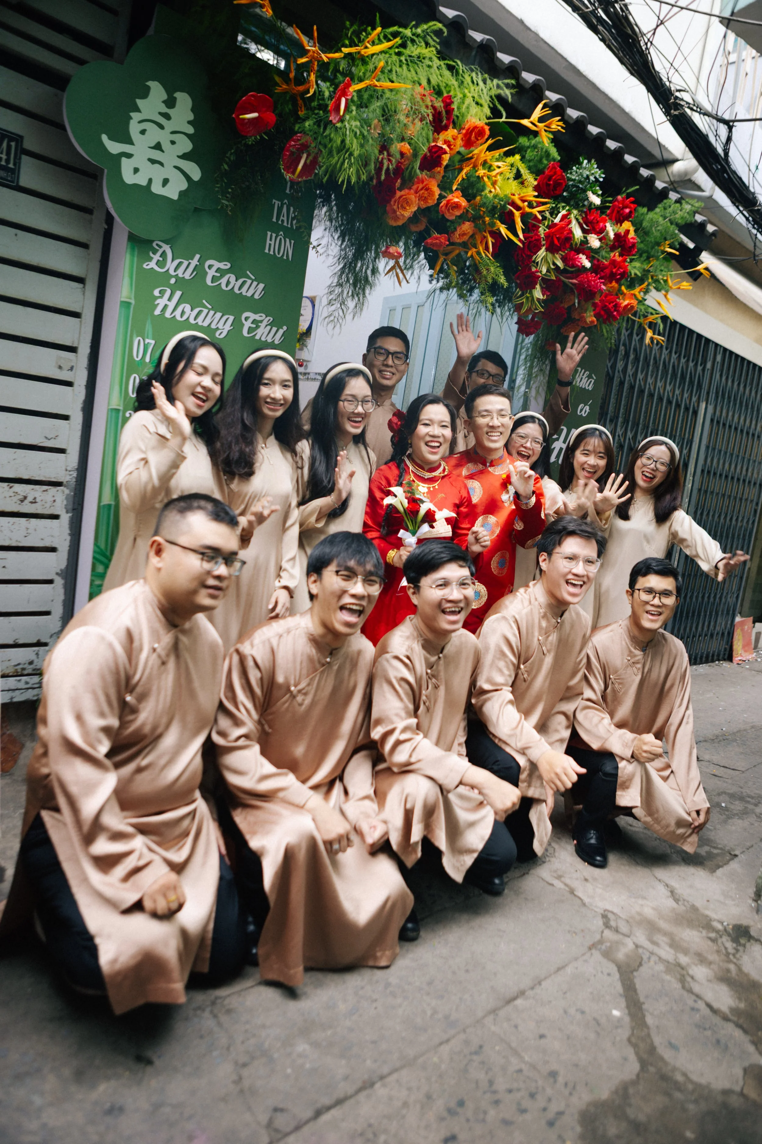 A group of people dressed in traditional Vietnamese clothing celebrating a wedding, standing in front of a floral arch and smiling for the photo.