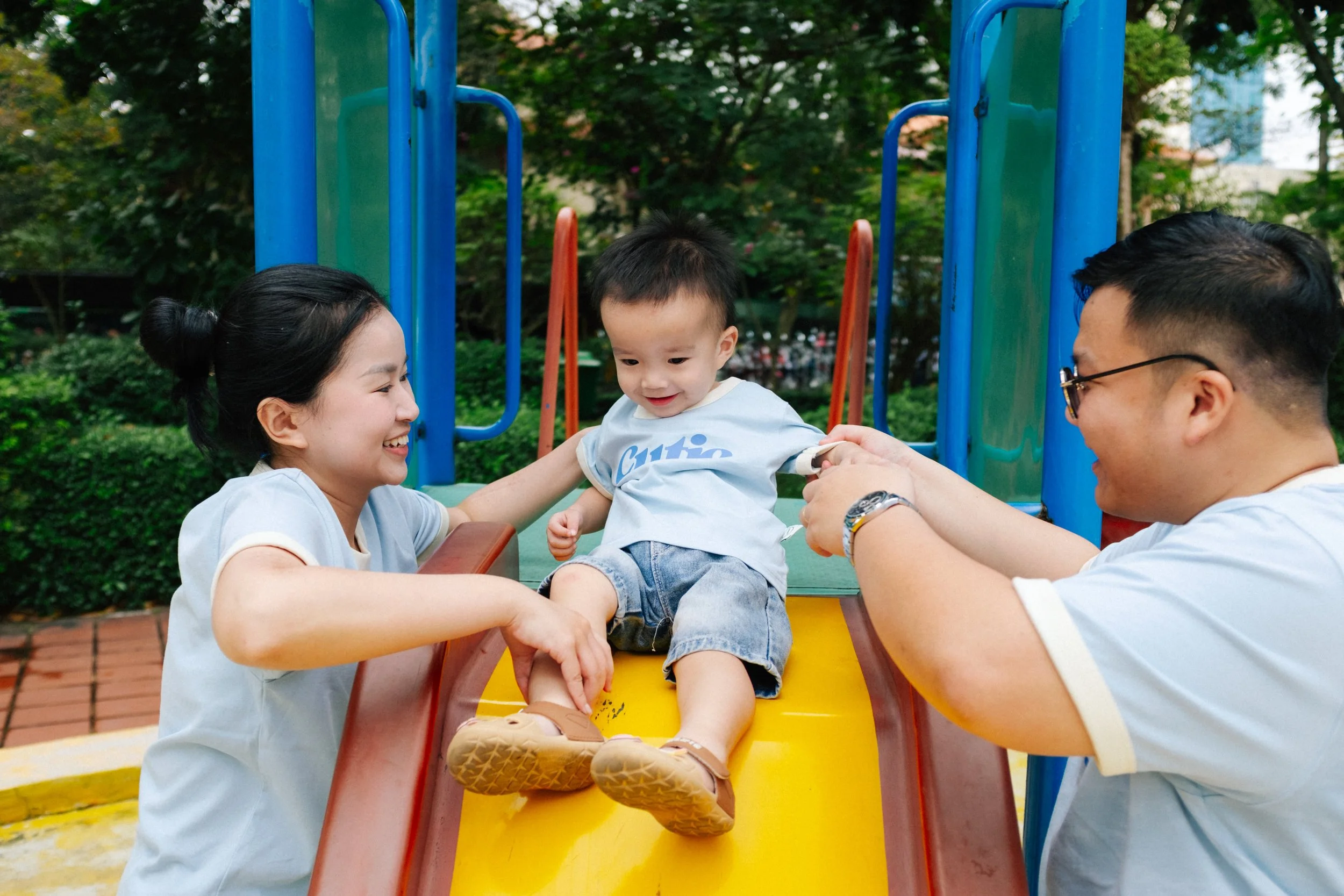 A young boy sitting at the top of a slide, smiling while holding hands with a woman on his left and a man on his right, both assisting him to slide down at a playground surrounded by greenery.