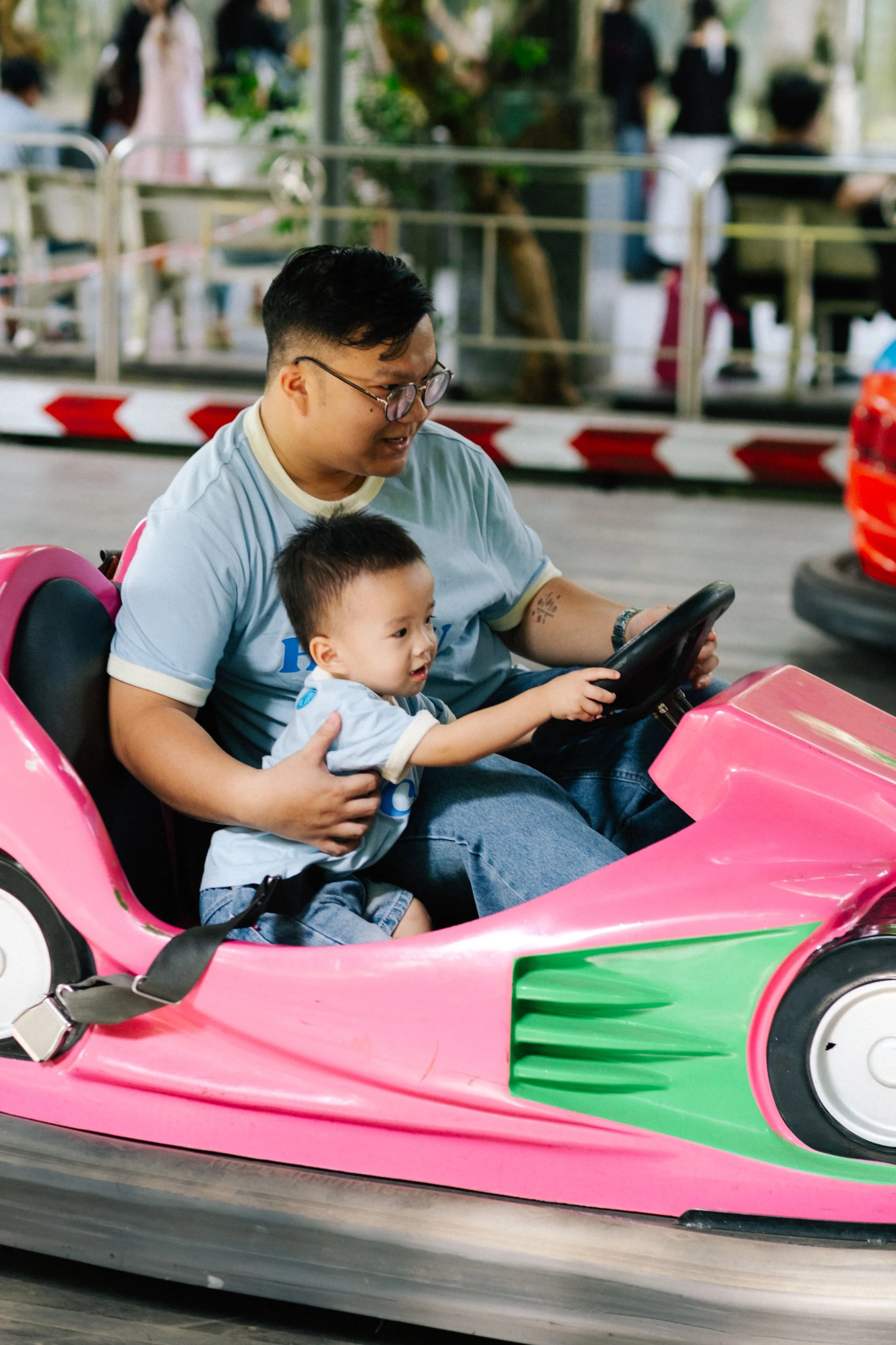 A smiling man and a young boy riding a pink bumper car at amusement park, with people in the background.