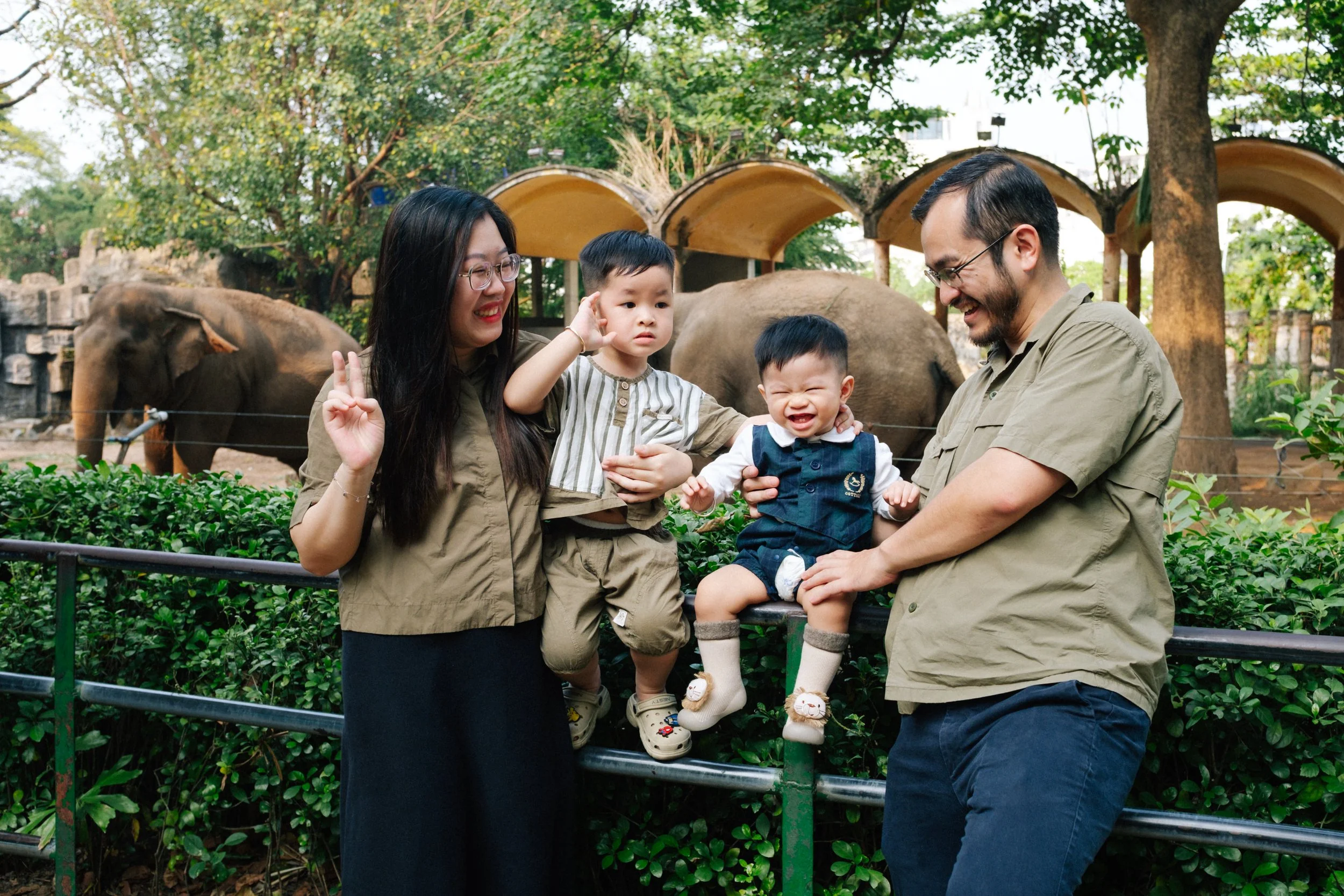 A family of four at an elephant enclosure in a zoo, with two children and two adults smiling and playing, surrounded by greenery and elephants in the background.