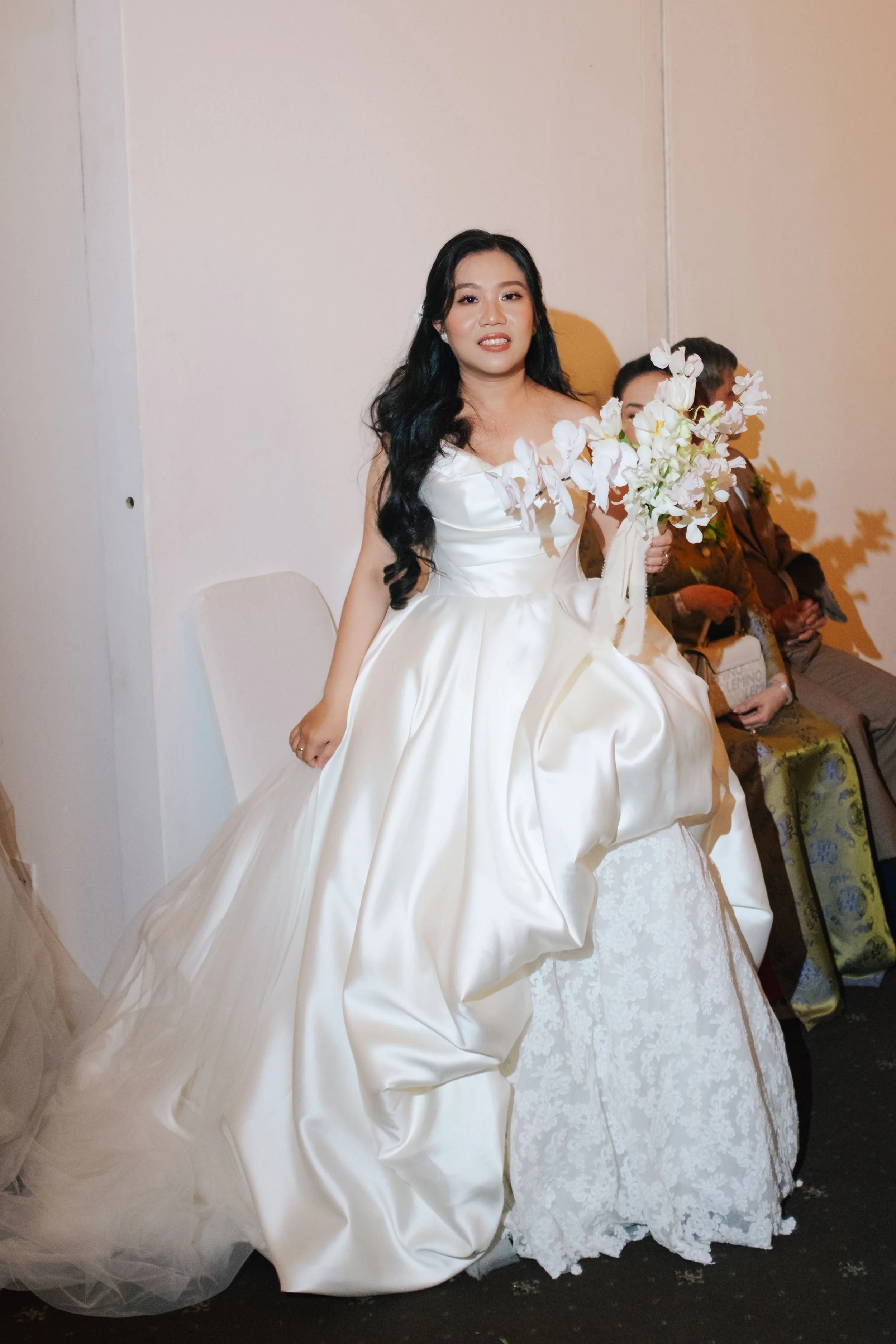 A woman in a wedding dress holding a bouquet of white flowers, sitting on a table at a wedding reception.