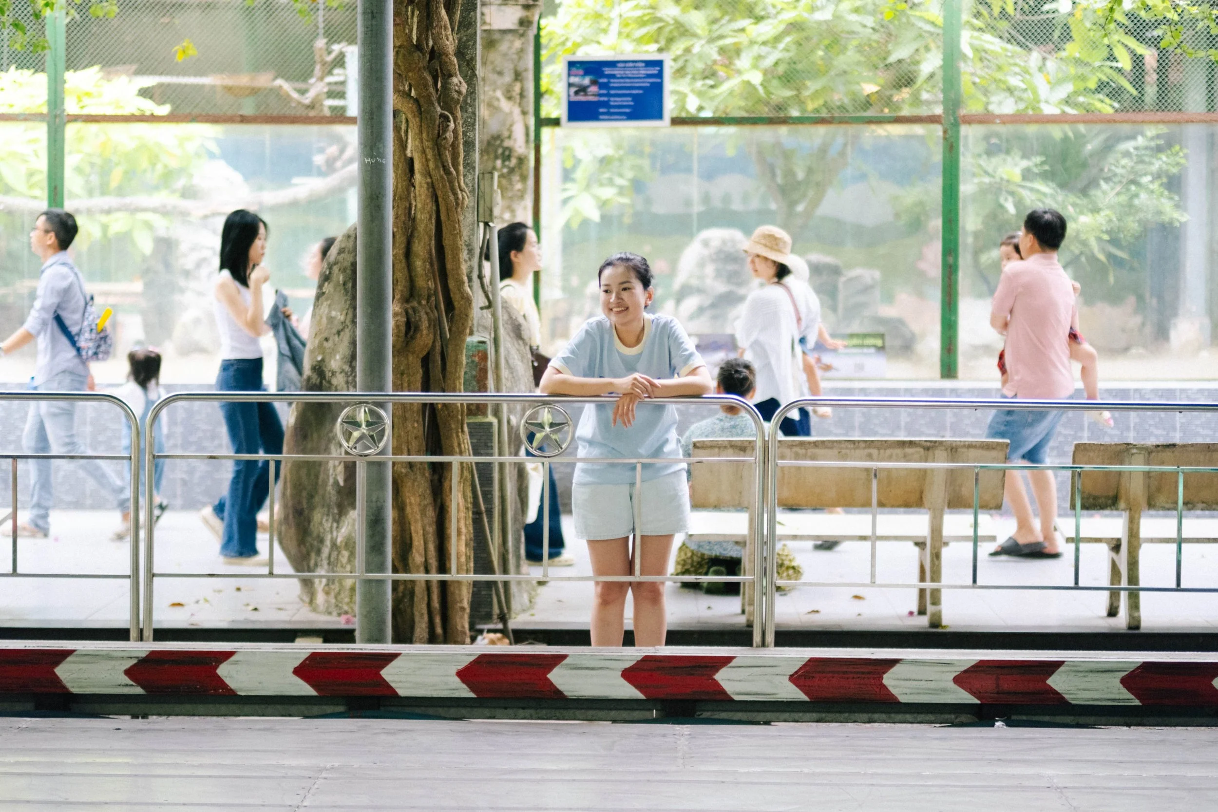 A young woman leaning on a metal railing inside an indoor zoo or botanical garden, with visitors walking past outside in a lush, green environment.