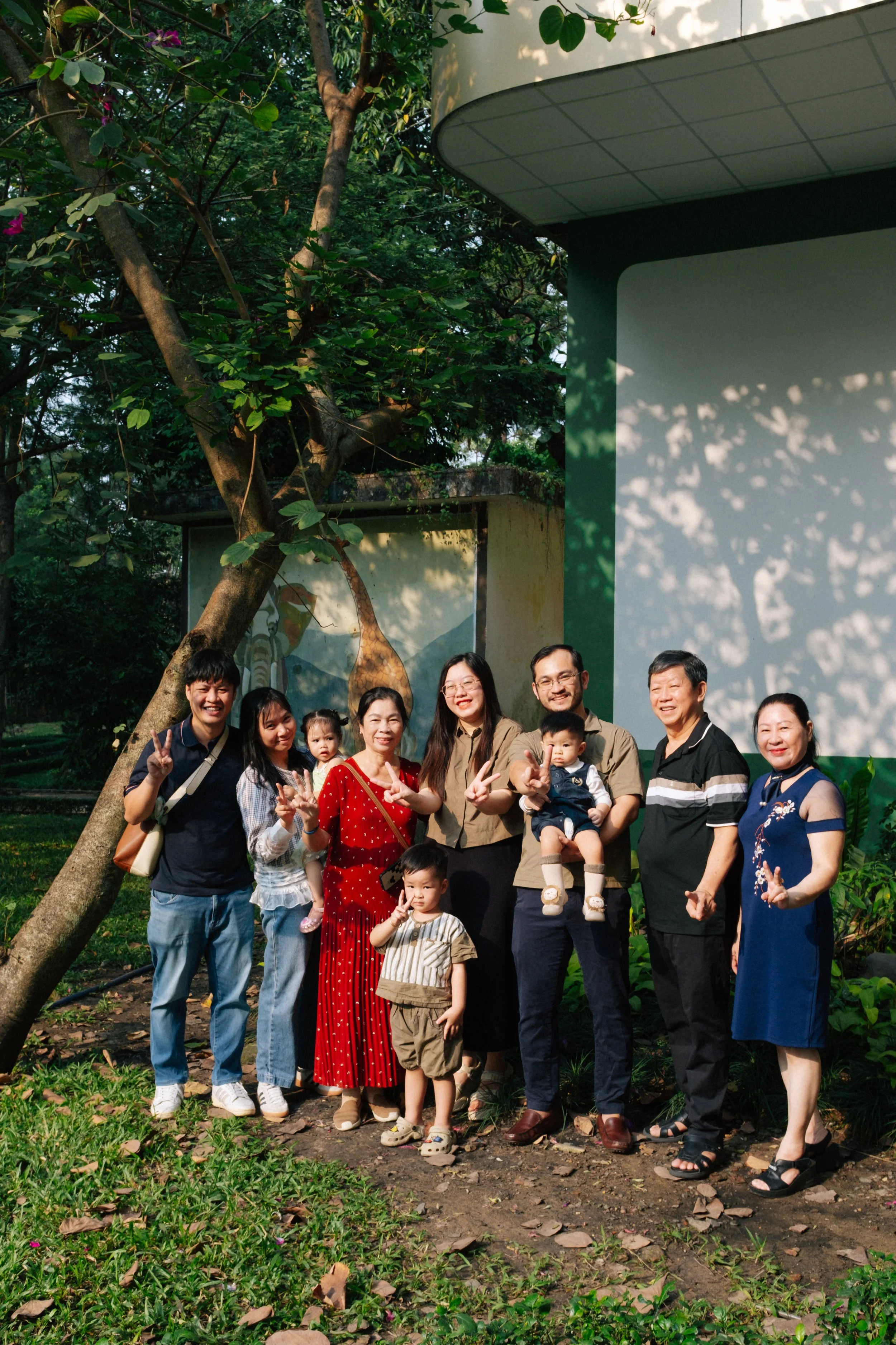 A group of people standing outdoors near a tree and a building, posing for a photo and making peace signs.
