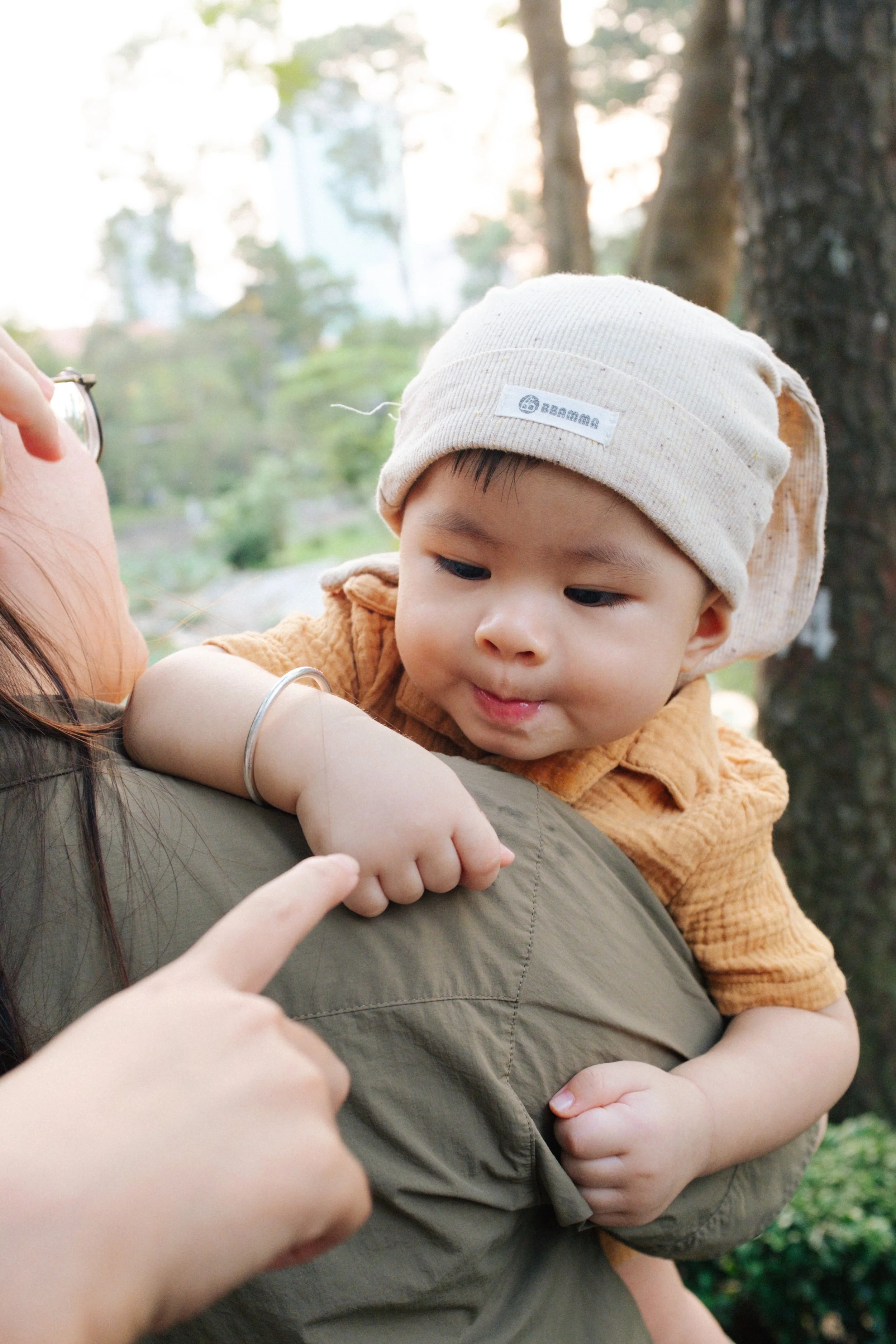 Child in a beige hat and orange shirt being held and gently touched by a woman, outdoors with trees and blurred background.