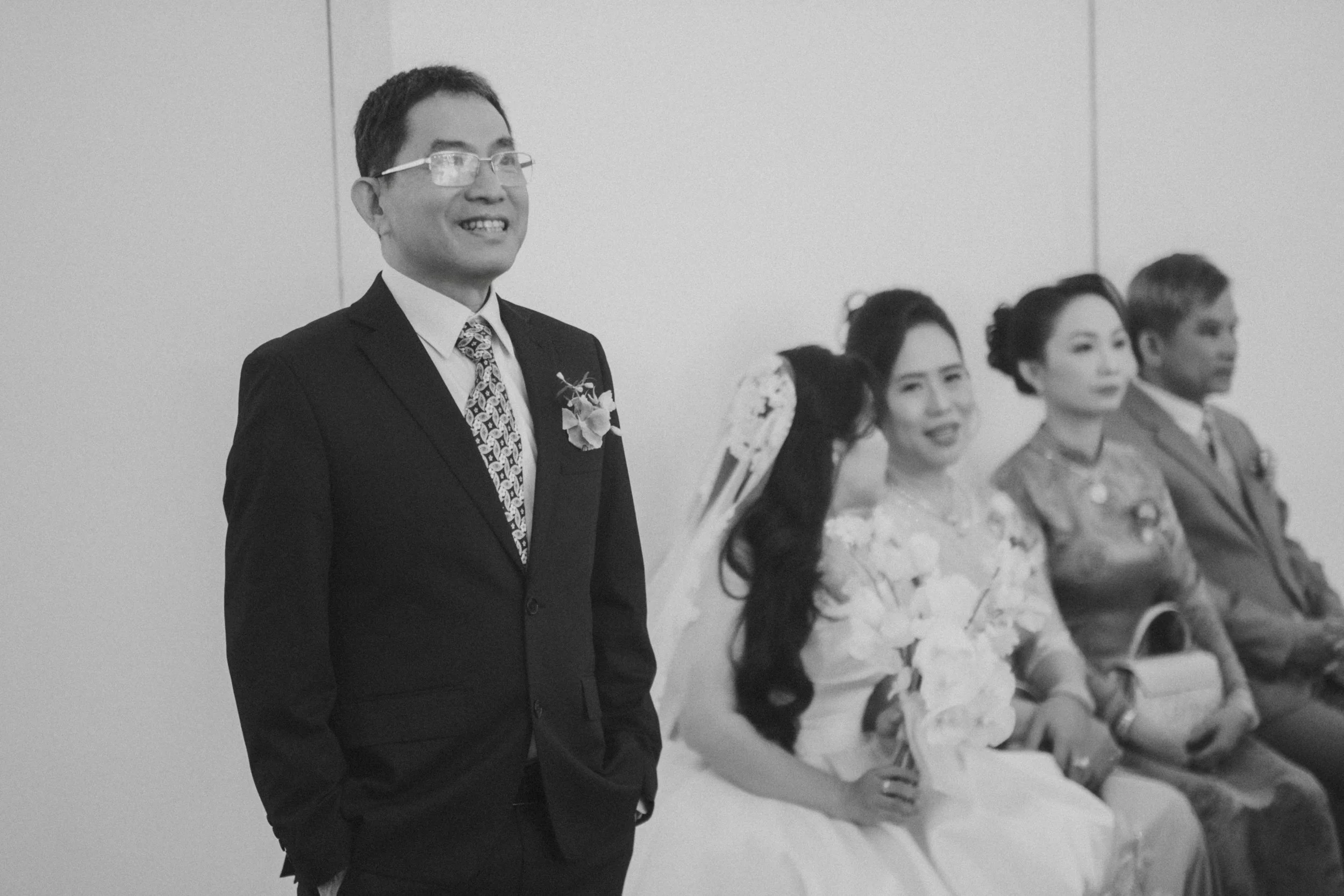 A black-and-white photo of a groom in a suit and glasses smiling, standing beside seated women dressed in traditional attire at a wedding ceremony.