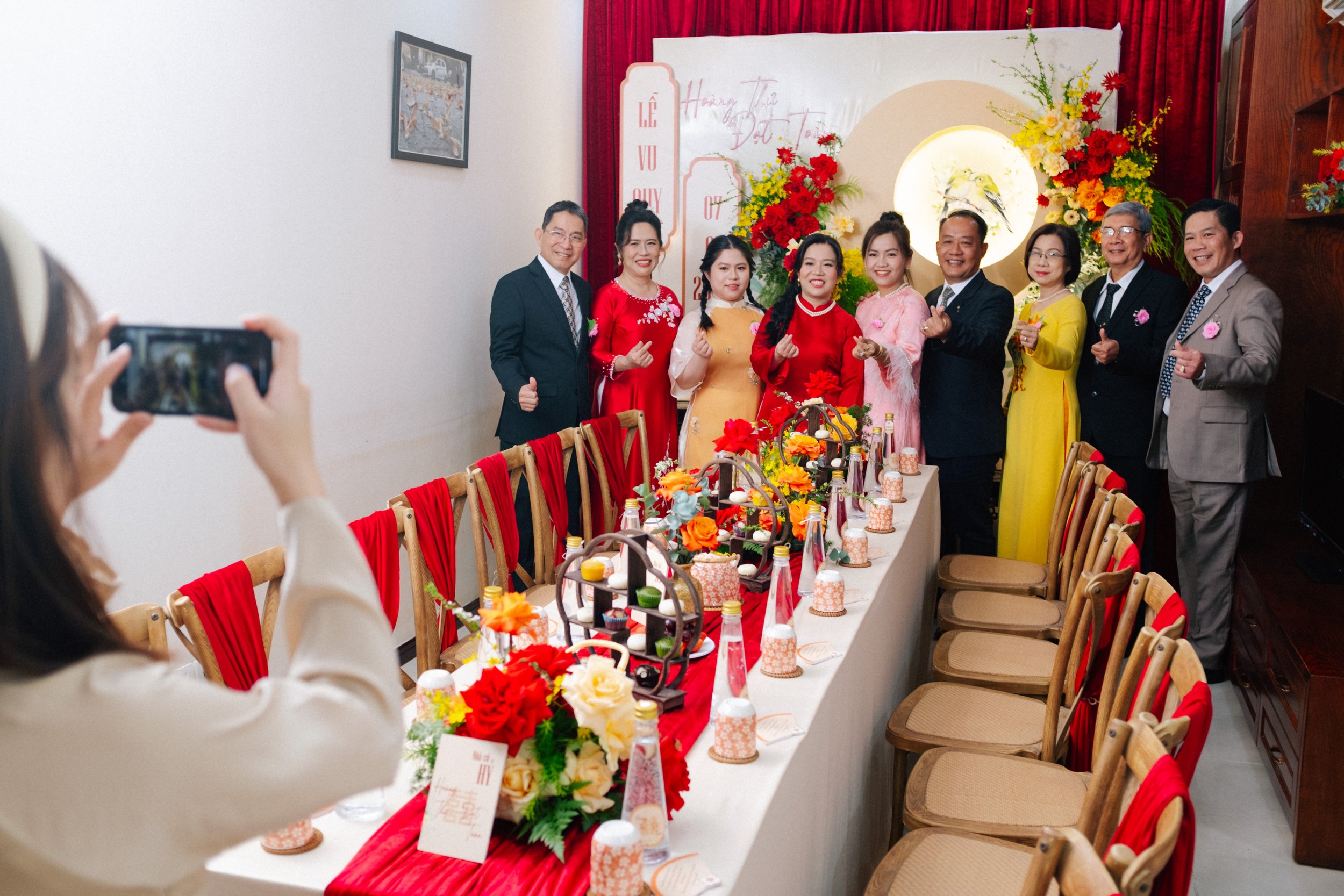 People posing for a photo at a wedding celebration, standing behind a decorated table with flowers, drinks, and desserts, with a woman taking their picture.