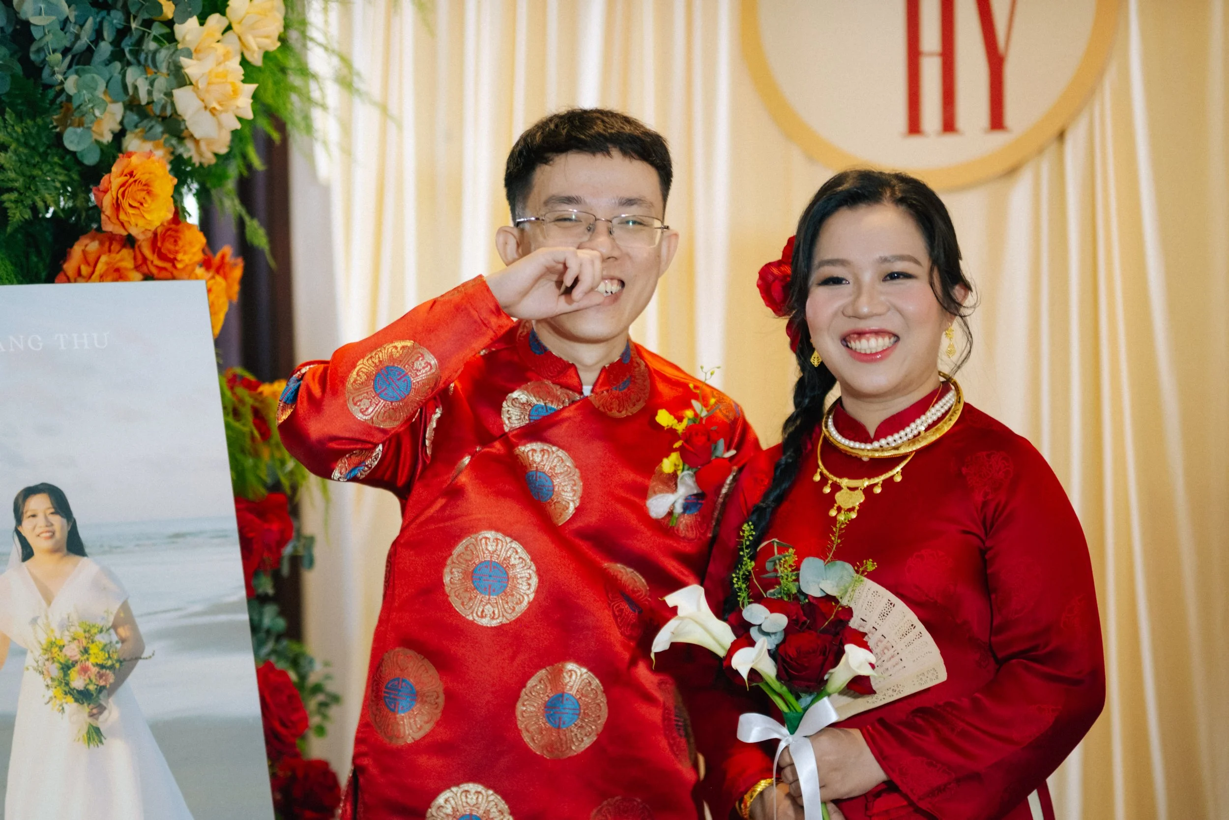 Couple in traditional red wedding attire smiling at the camera, woman holding a bouquet of flowers, background decorated with flowers and curtains.