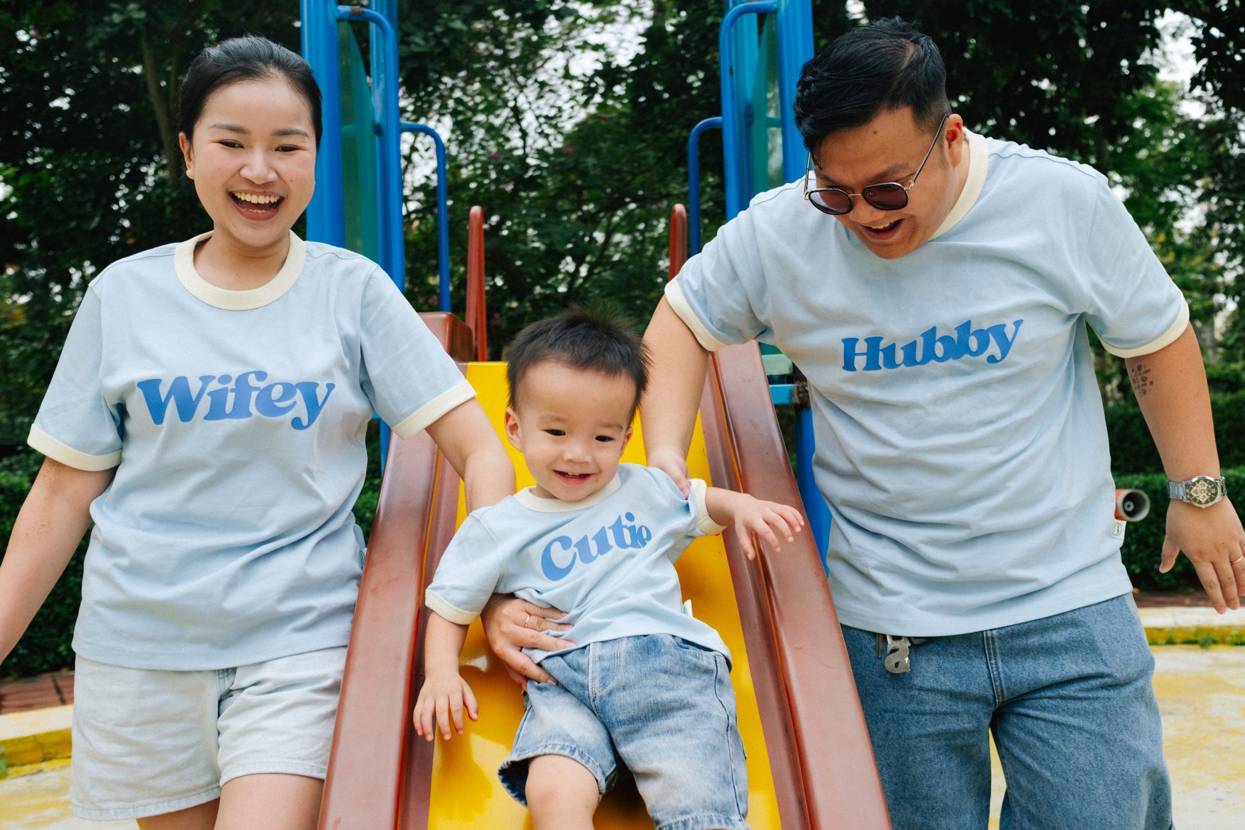 A family of three, including a woman, a man, and a young boy, is smiling and playing on a slide at a park. They are all wearing matching T-shirts with words on them, the woman’s reads "Wifey," the man’s reads "Hubby," and the boy’s reads "Cutie."