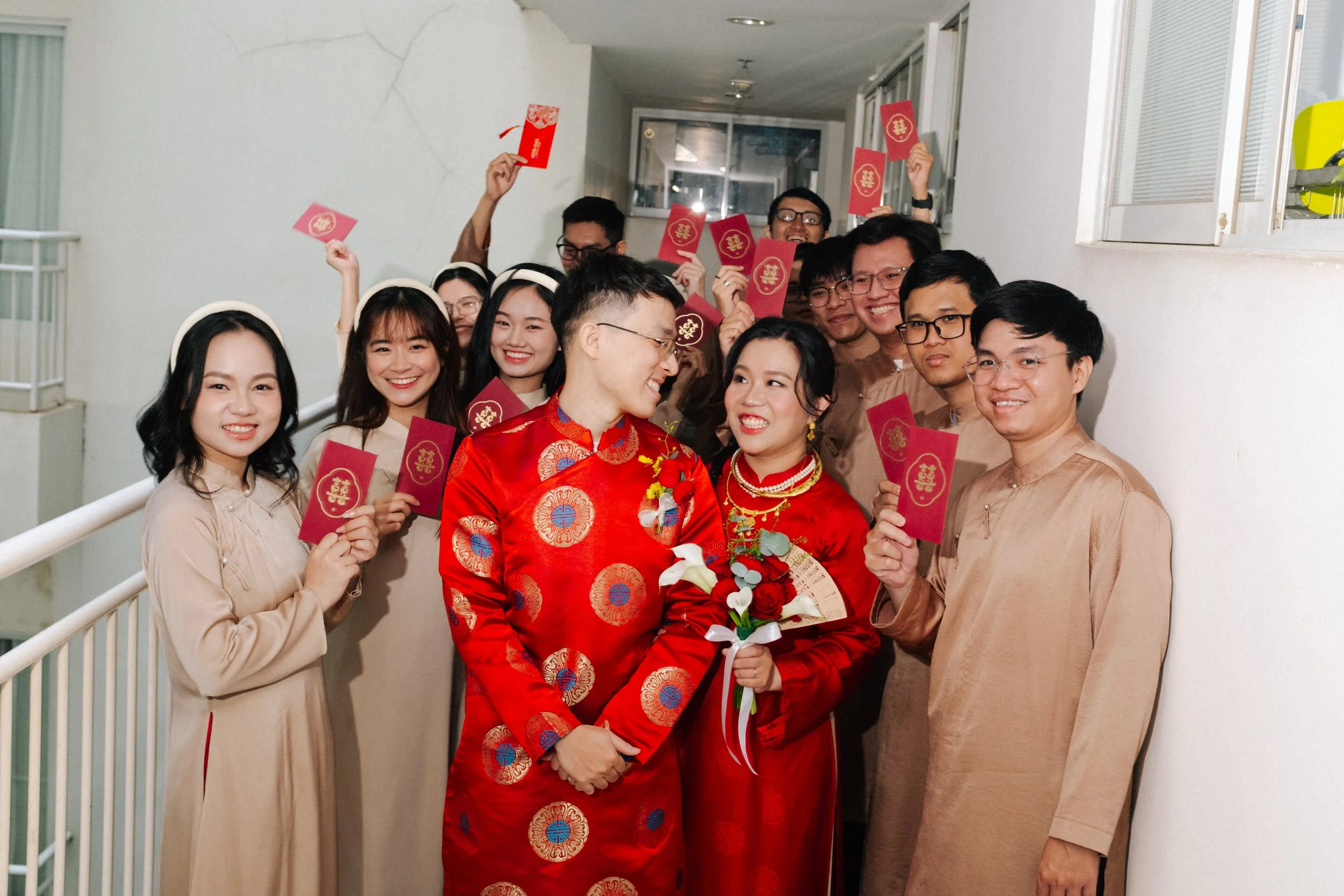 A group of people gathered in celebration, with a couple in traditional red Vietnamese attire at the center, holding flowers and smiling at each other. Others are holding red envelopes, indicating a festive occasion, likely a wedding or Lunar New Yea