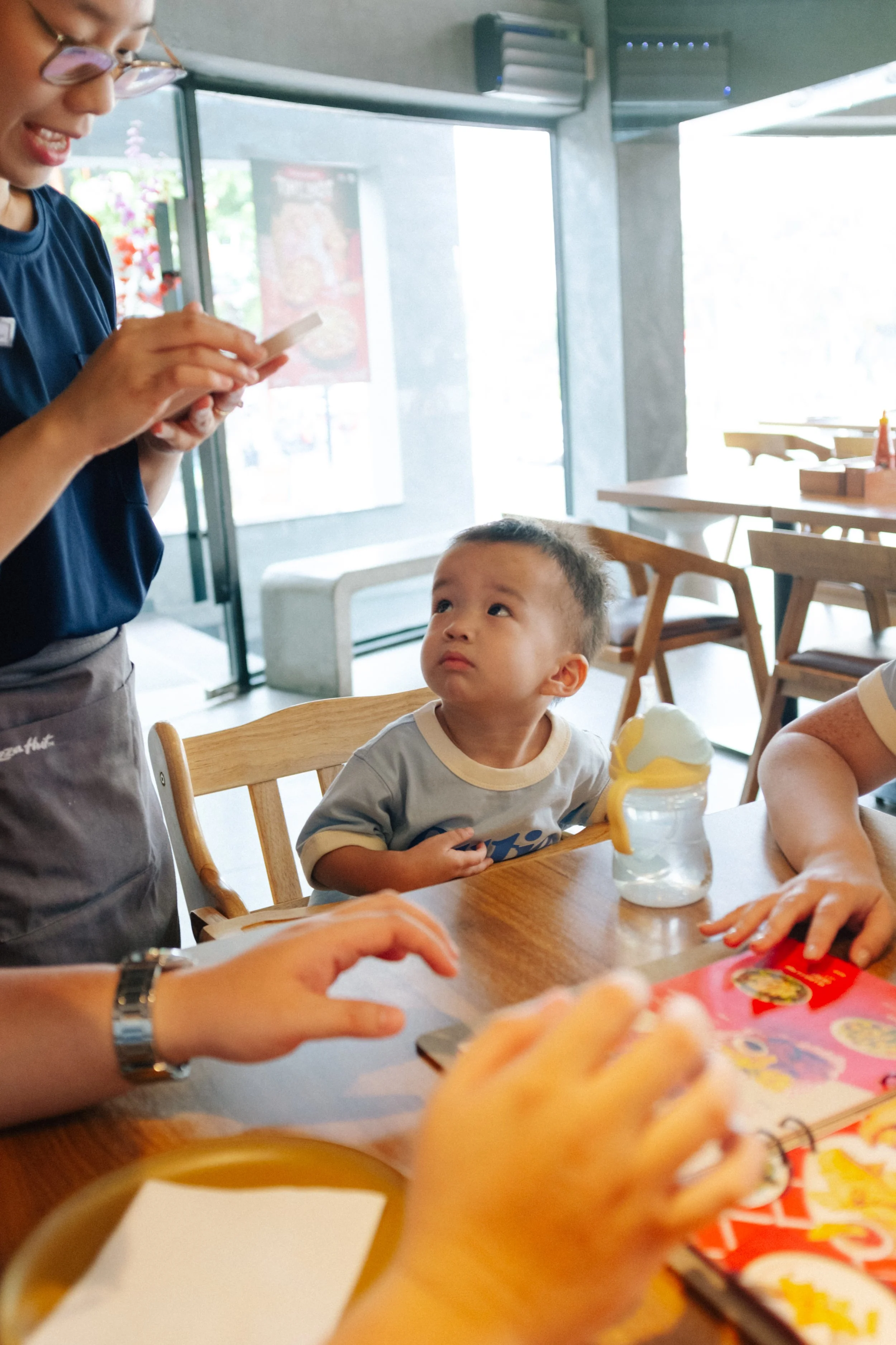 A young boy sitting at a wooden table in a restaurant, looking up at a woman who is standing and using her phone. Other people are also seated at the table, with a water bottle and a colorful menu in front of them.