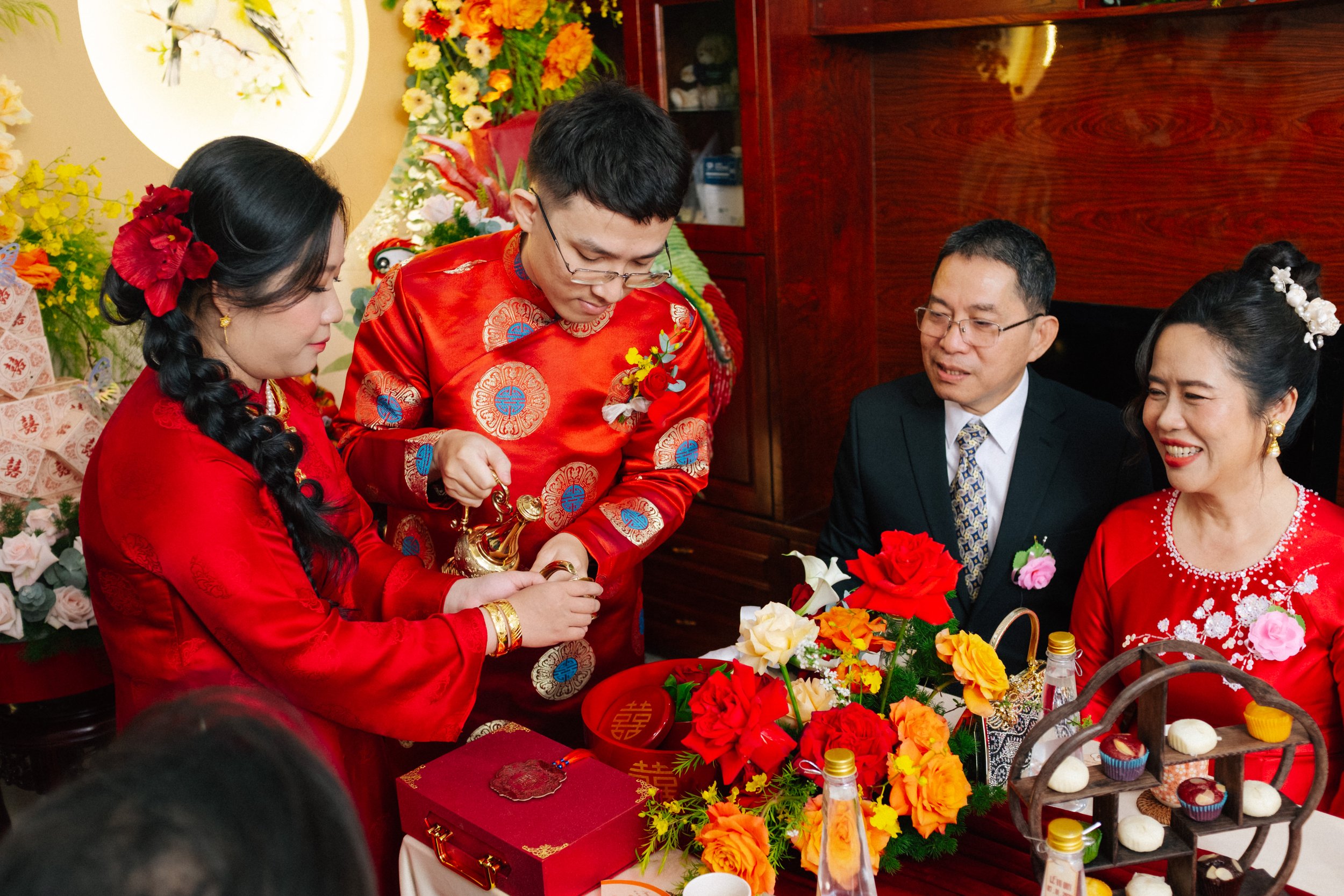 A traditional Asian wedding tea ceremony with family members dressed in red, pouring tea and exchanging gifts, surrounded by floral decorations, on a decorated table.