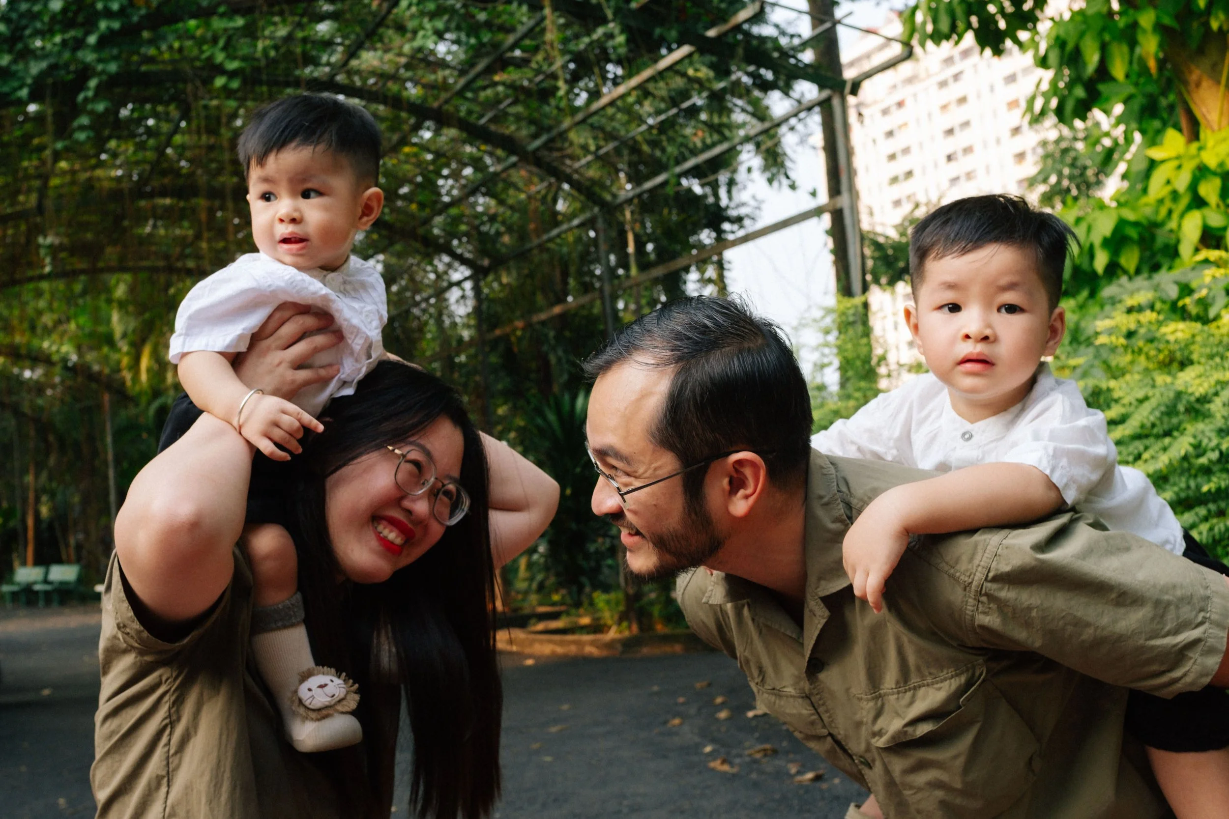 A family of four with two young boys playing outdoors in a park, with trees and greenery in the background. The woman is holding one boy on her shoulders, and the man has the other boy on his back. They are all smiling and looking at each other.
