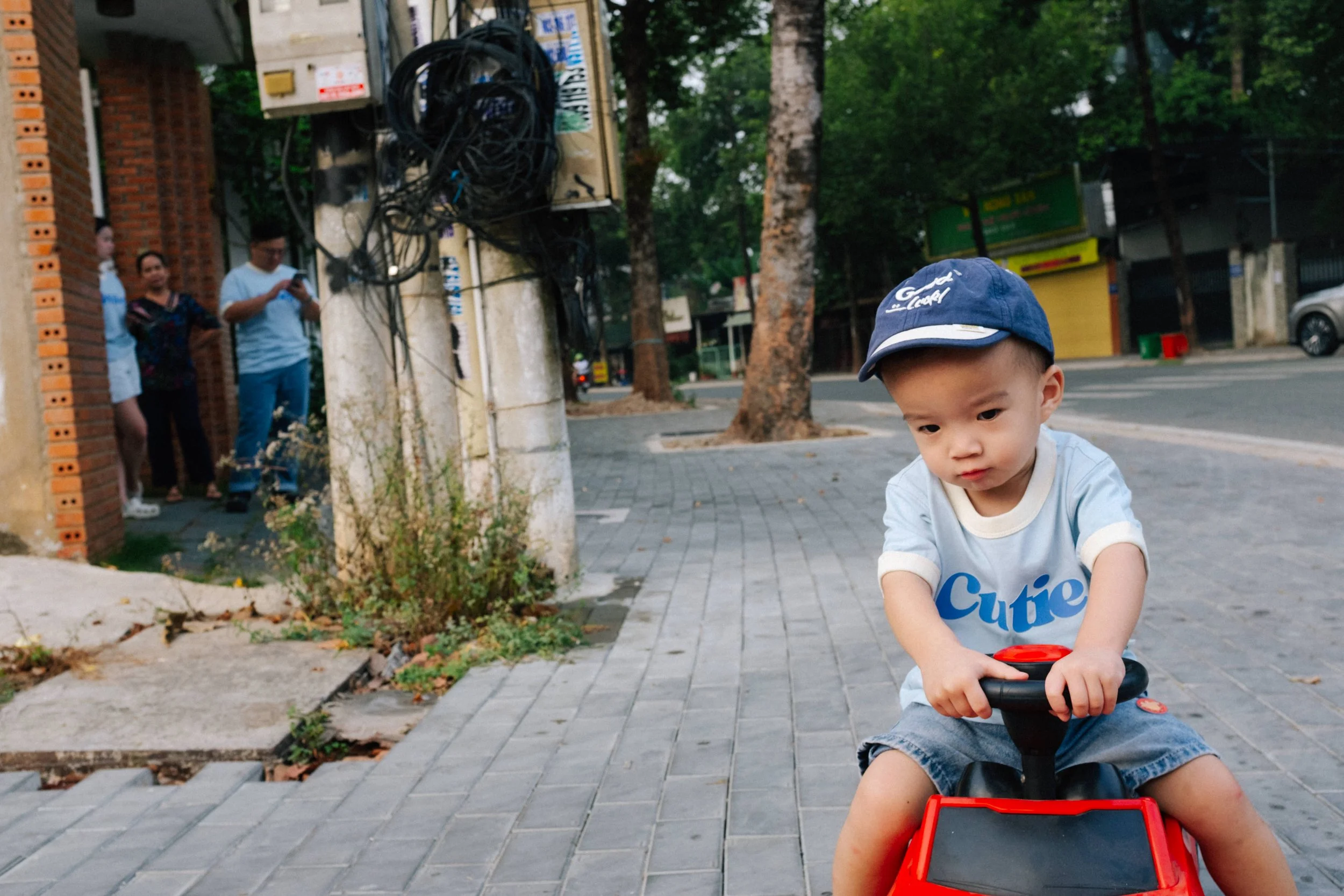 A young boy wearing a blue baseball cap and a gray T-shirt with the word 'Cutie' on it, riding a red toy car on a sidewalk.
