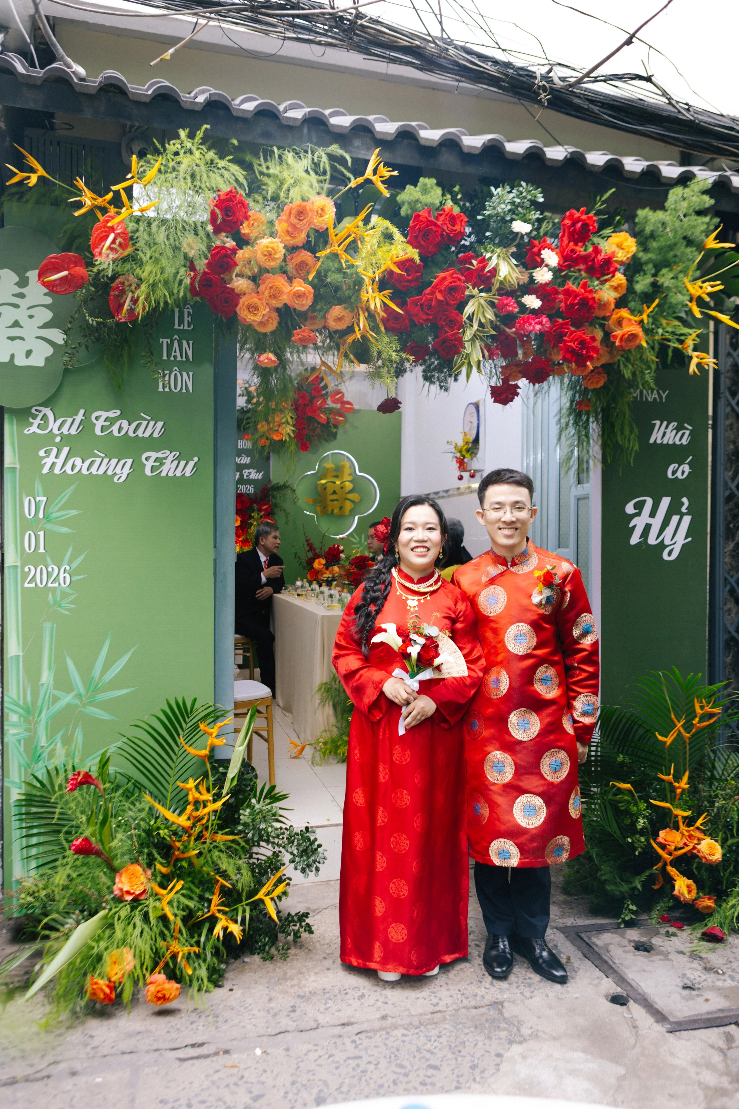 A couple dressed in traditional Vietnamese red áo dài, holding a bouquet, standing in front of a decorated entrance with green banners and vibrant red, orange, and yellow flowers, celebrating a special occasion.