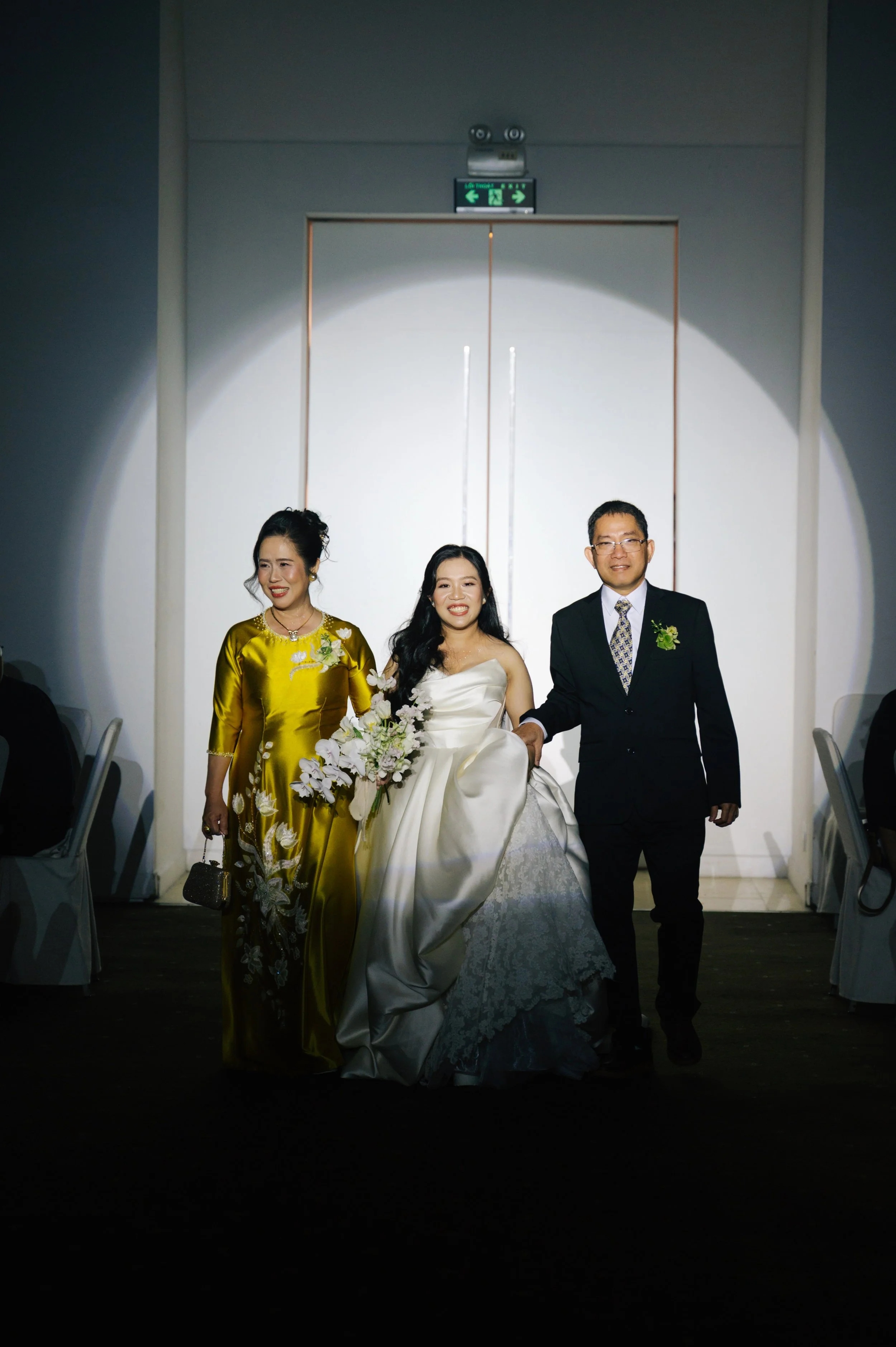 A bride in a white wedding gown holding a bouquet, walking with her parents at a wedding reception. The bride is smiling while her mother, in a gold dress, and her father, in a black suit, walk alongside her. They are under a spotlight in a banquet h