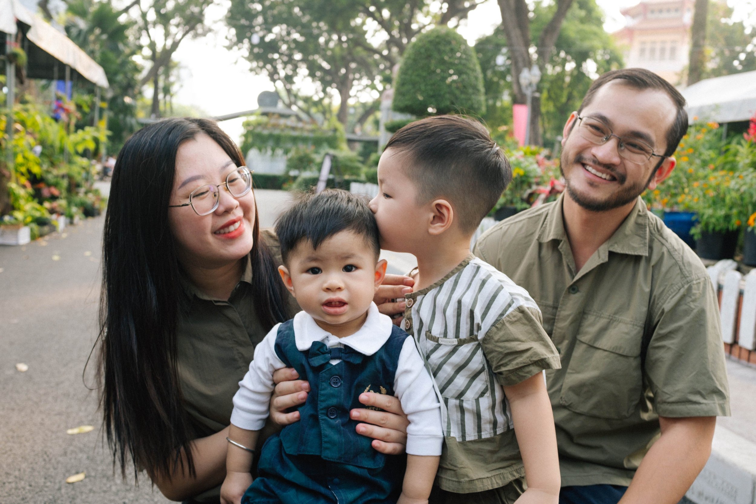 A happy family of four outdoors, with a woman, a man, and two young boys, one kissing the other on the head, surrounded by trees and plants.
