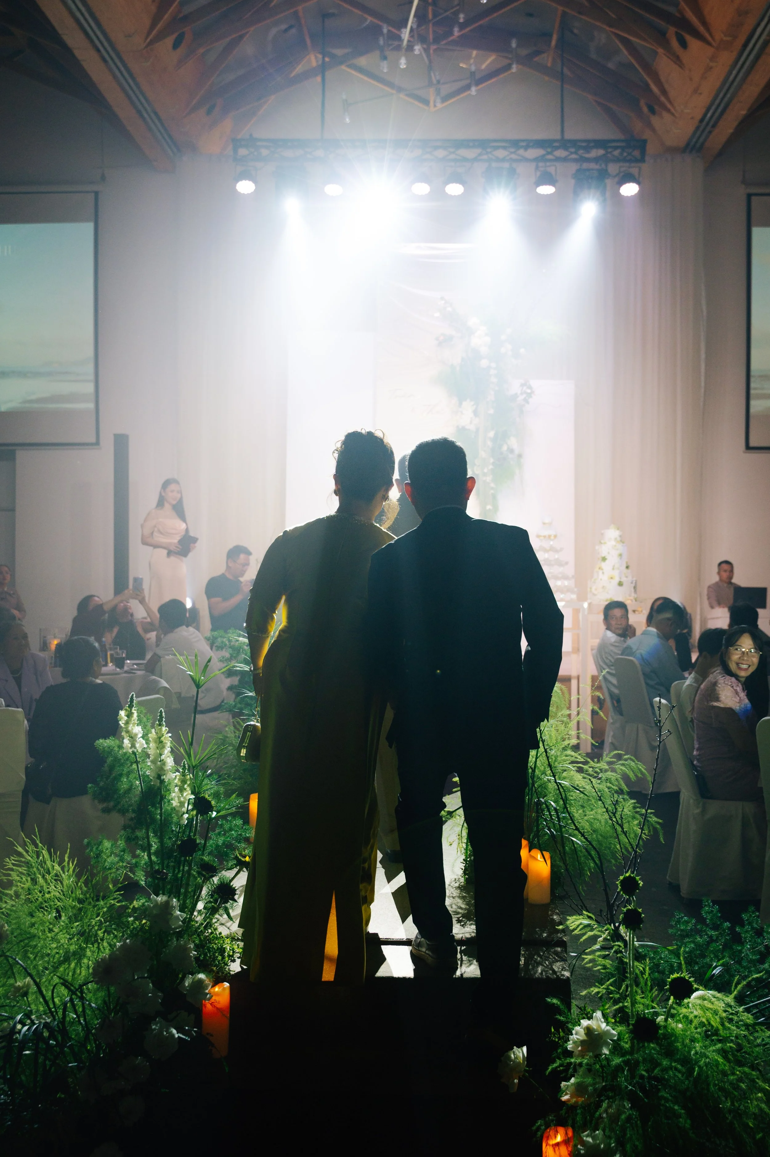 Silhouetted couple stands together at a wedding reception, overlooking the dance floor, with bright stage lights shining down and guests seated at tables.